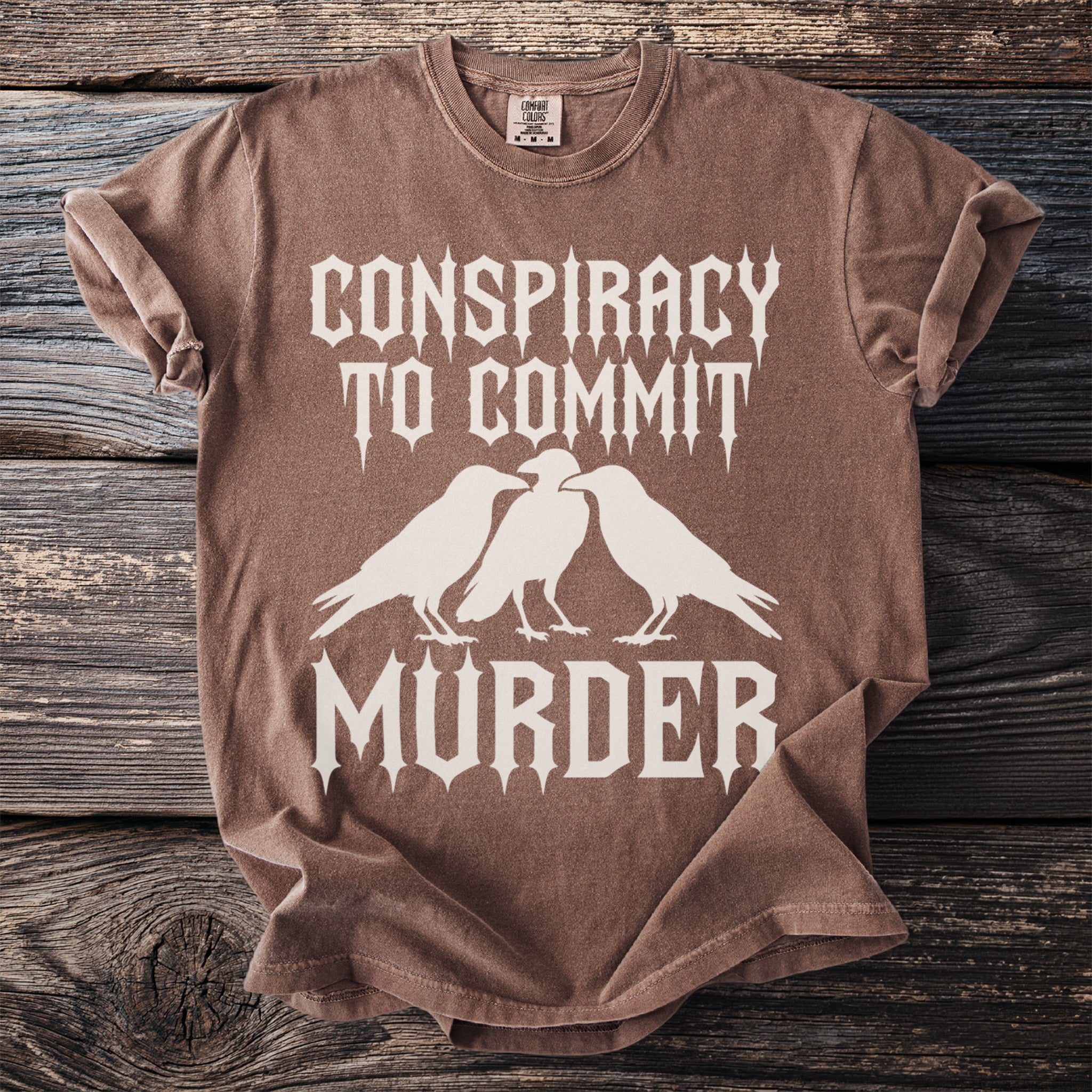 Brown t-shirt with white text and three ravens: "Conspiracy to Commit Murder".