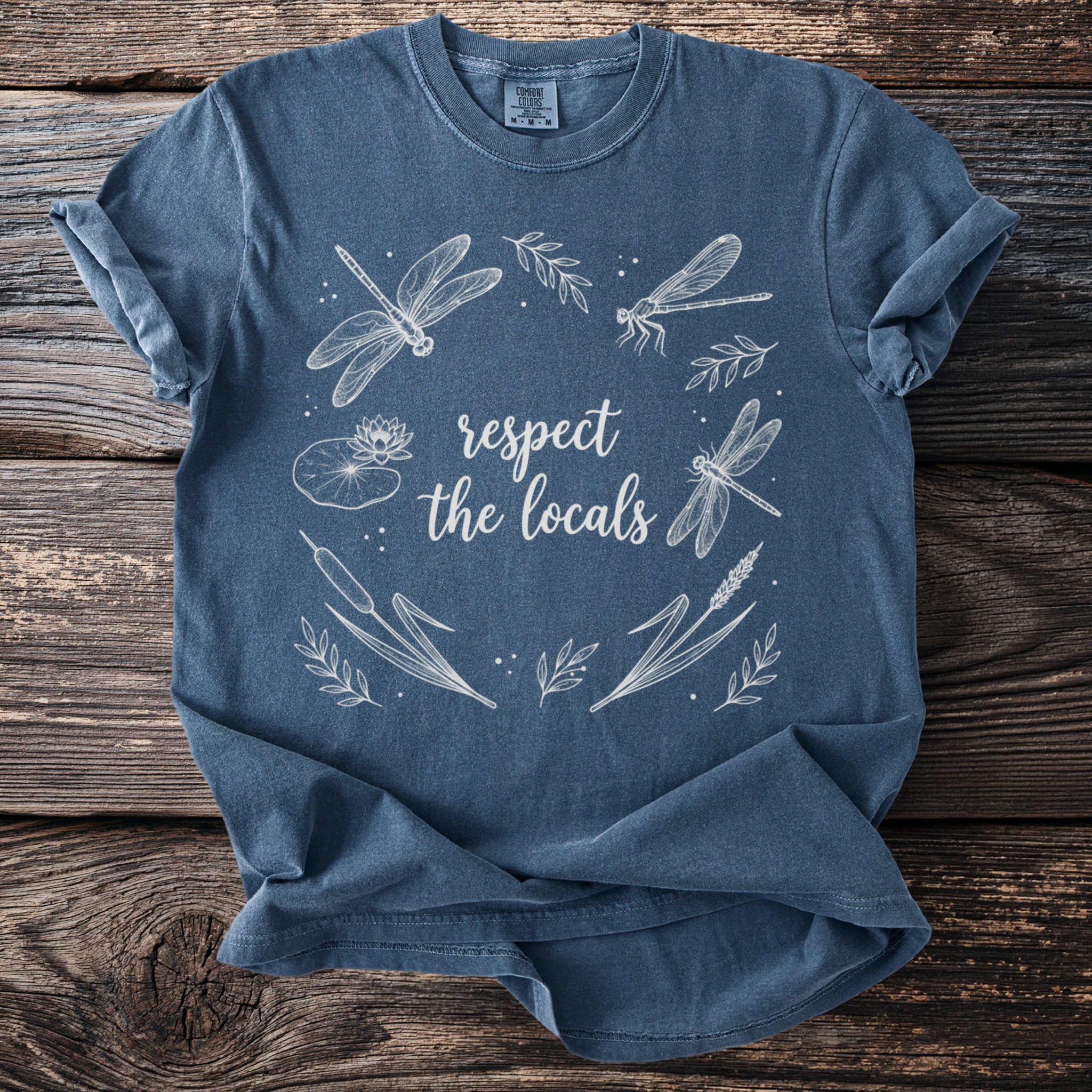 Blue t-shirt with white line art of dragonflies, water lilies, and reeds with the text "respect the locals".