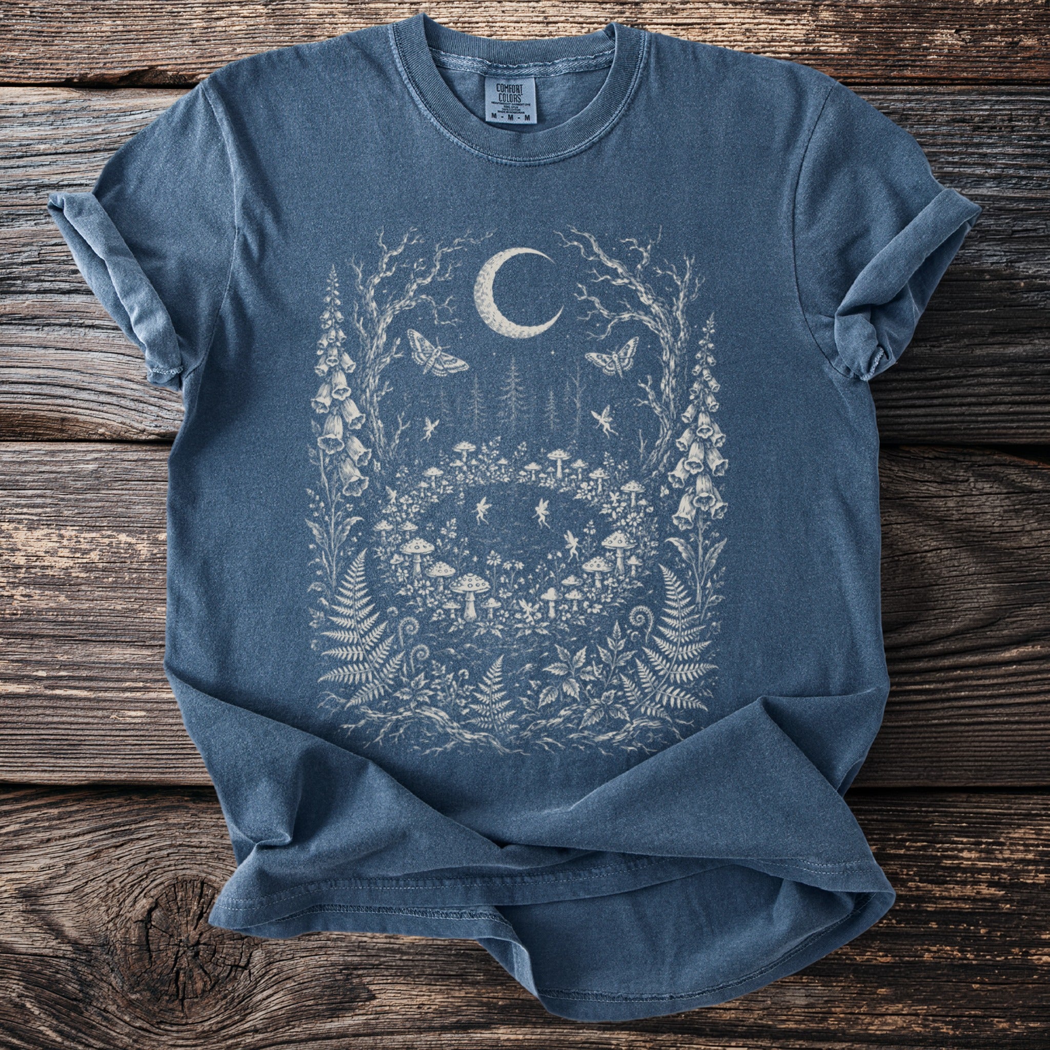 Blue t-shirt with a mystical forest scene featuring a moon, moths, mushrooms, and fairies.