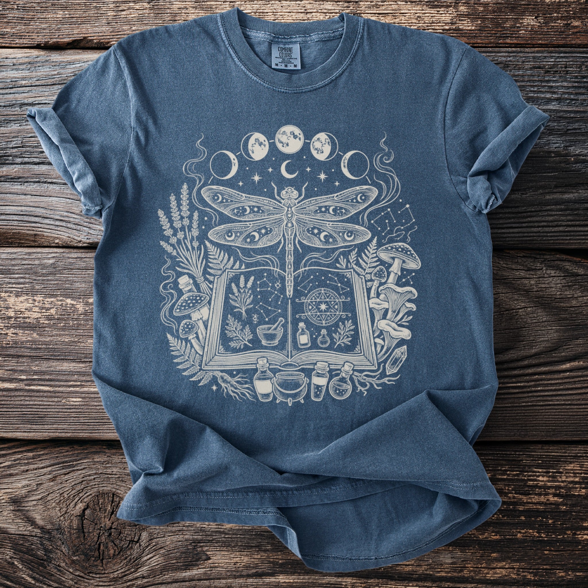 Blue t-shirt with a white graphic of a dragonfly over an open book and mystical elements.