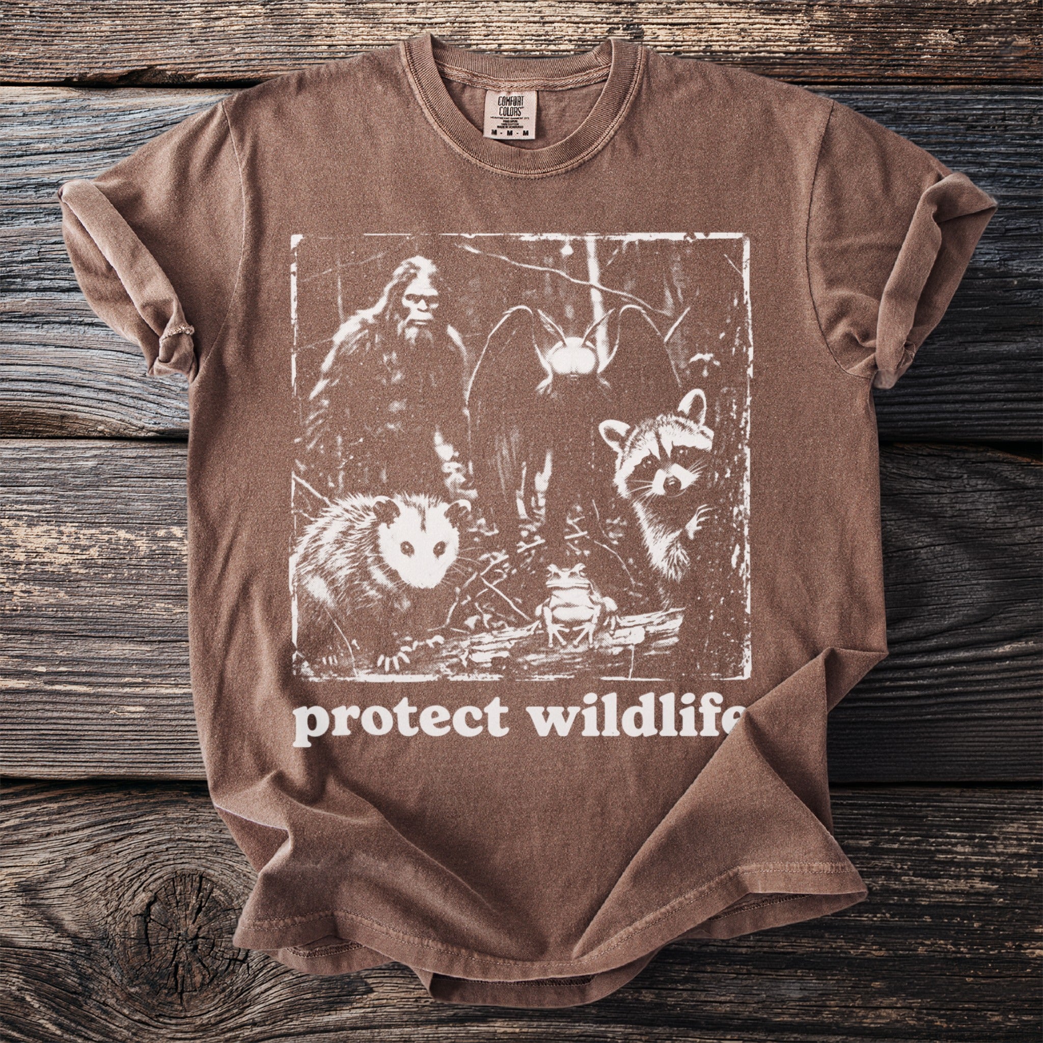 Brown t-shirt with graphic of Bigfoot, owl, raccoon, opossum, and frog with "protect wildlife" text.