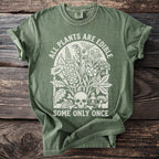 Green t-shirt with white graphic of plants, skull, bones, and text: "ALL PLANTS ARE EDIBLE SOME ONLY ONCE".