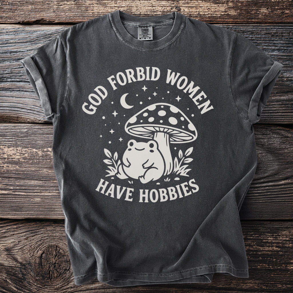 Dark grey t-shirt with "God forbid women have hobbies" text and frog under mushroom graphic.