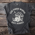 Dark grey t-shirt with "God forbid women have hobbies" text and frog under mushroom graphic.