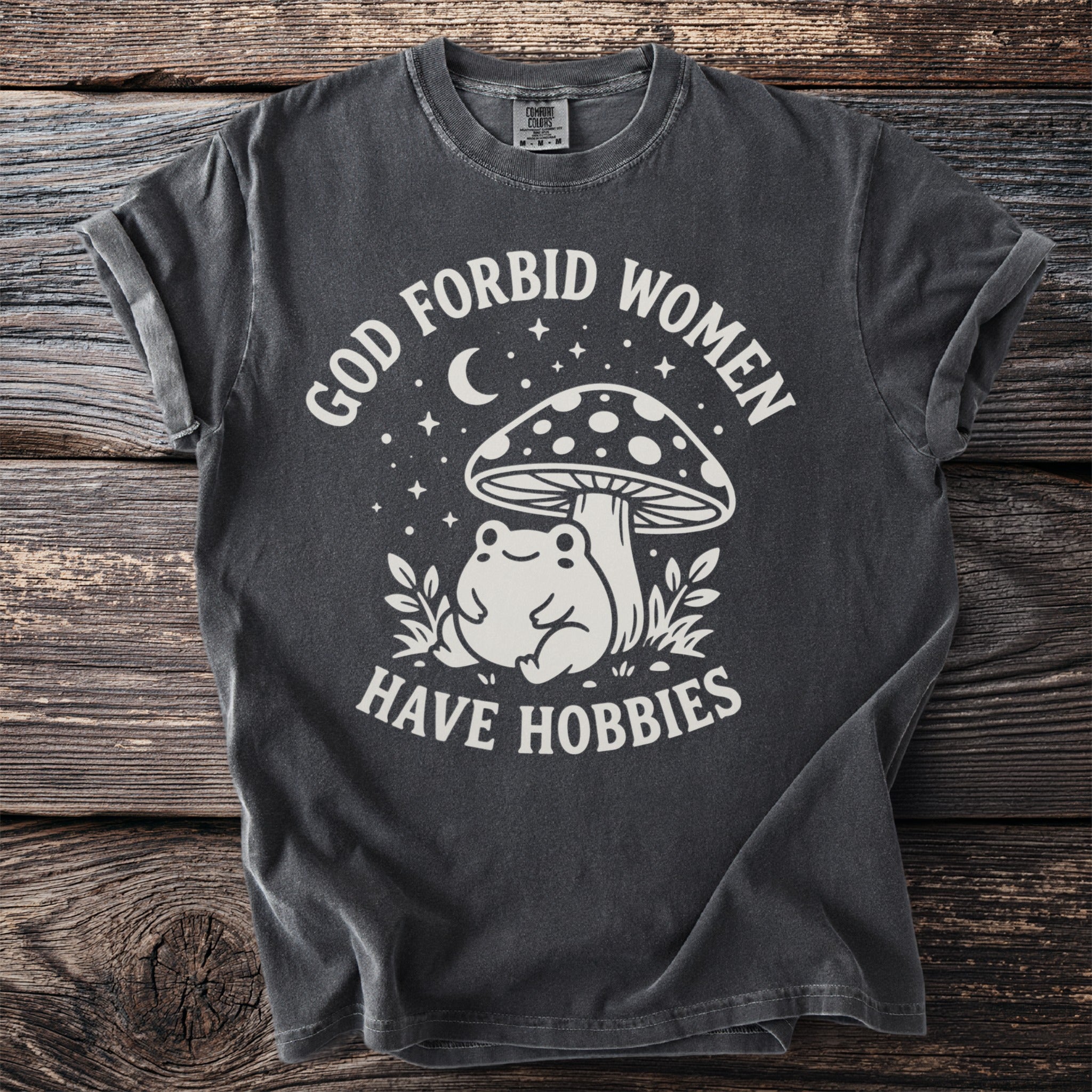 Dark grey t-shirt with "God forbid women have hobbies" text and frog under mushroom graphic.
