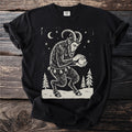 Black t-shirt with a cream folk-cut print: Krampus crouches among snowy pines, packing a snowball between his clawed hands.
