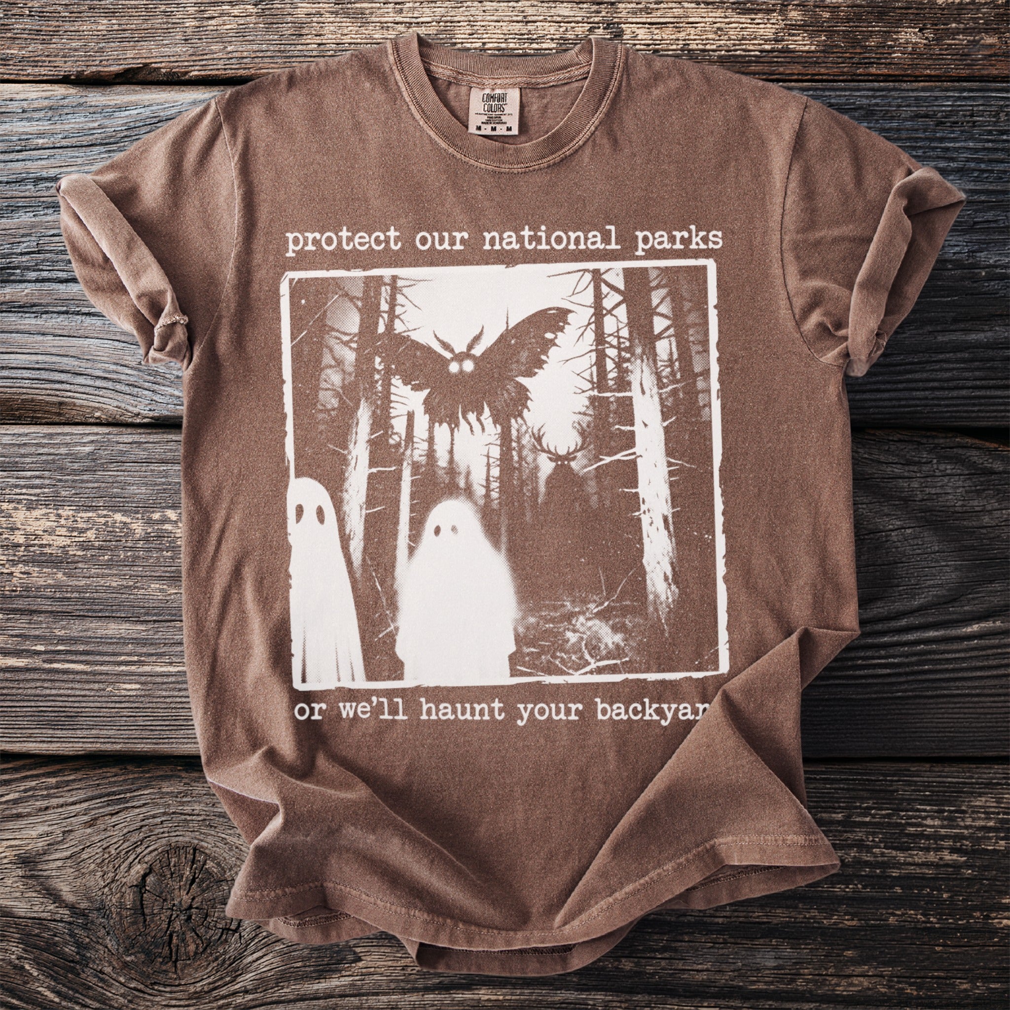 Brown t-shirt with "protect our national parks or we'll haunt your backyard" text and forest graphic.