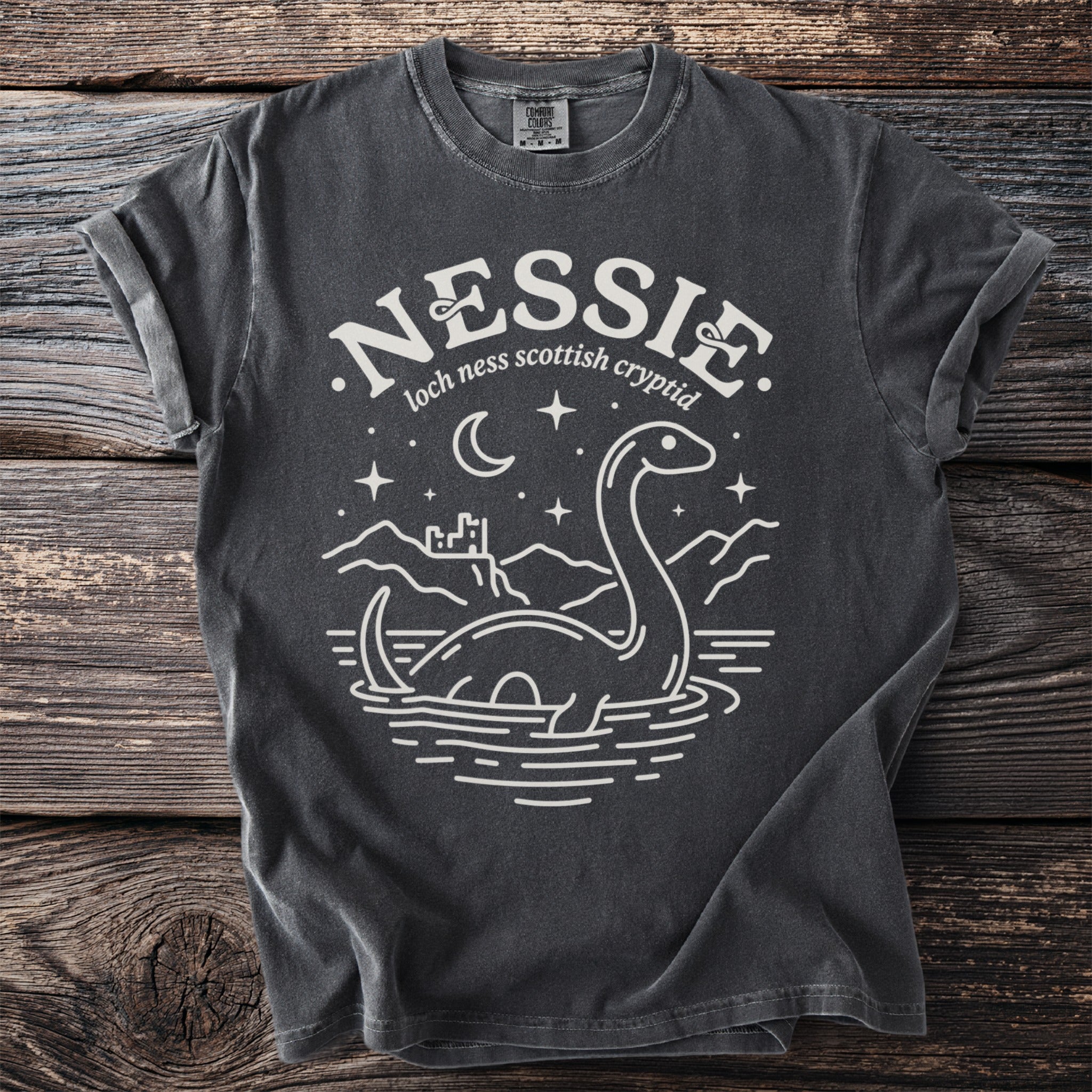Dark gray t-shirt with white graphic of Nessie, Loch Ness, castle on wooden background.