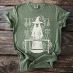 Green t-shirt with a white graphic of a witch, plants, jars, and bottles.