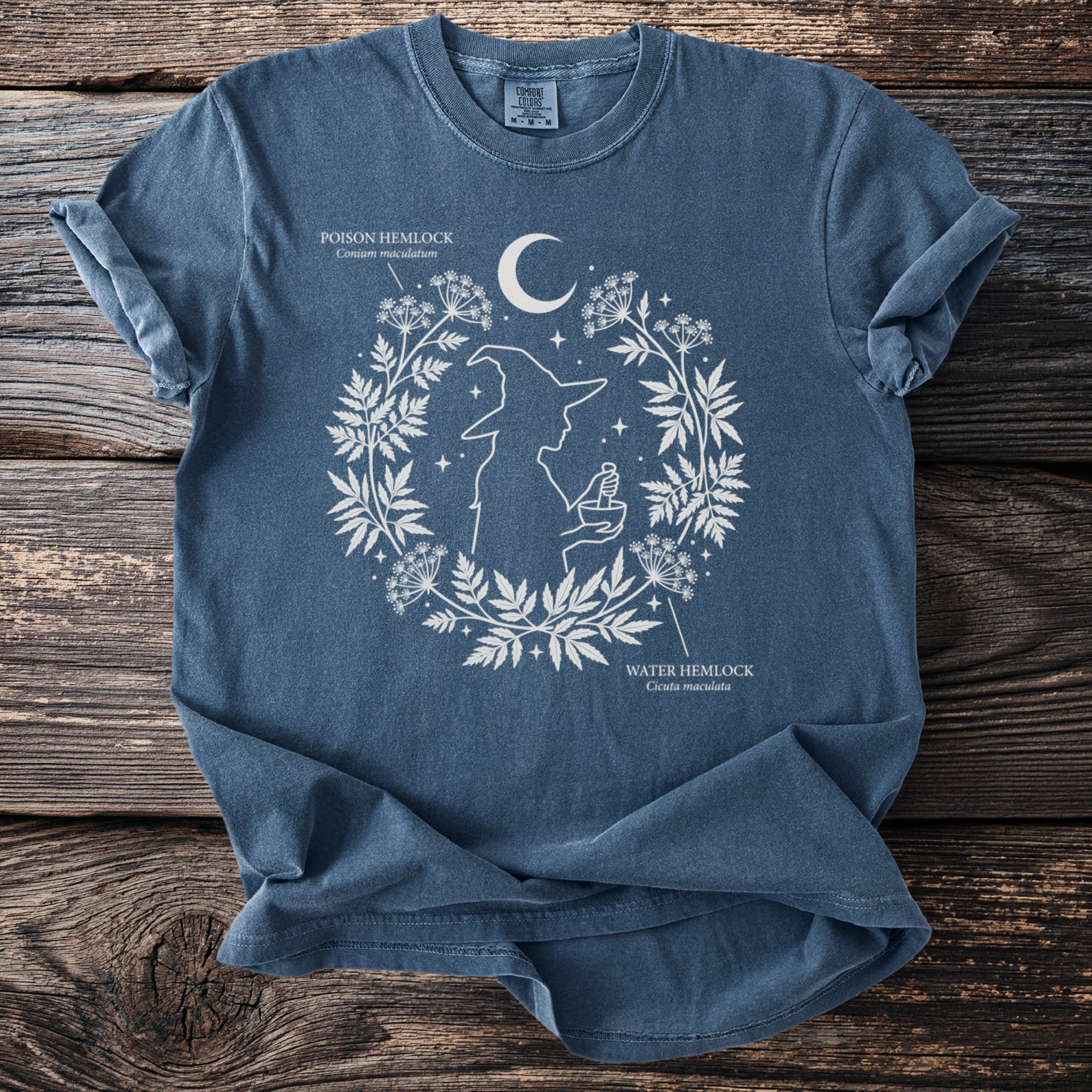 Blue t-shirt with white graphics of a witch silhouette and botanical designs, featuring text for poison and water hemlock.