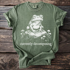 Green t-shirt with a white graphic of a frog on a rock surrounded by mushrooms and the words "currently decomposing".