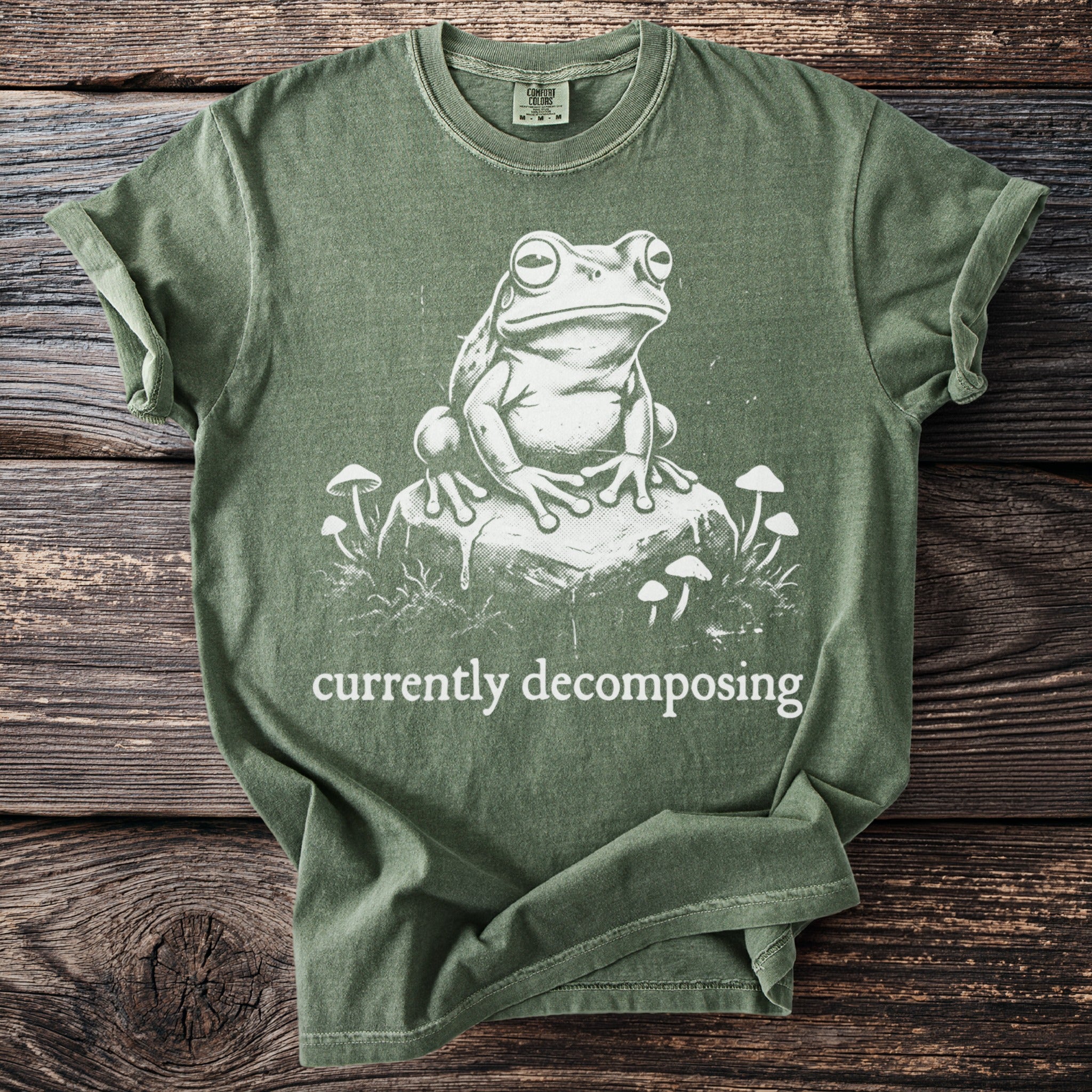 Green t-shirt with a white graphic of a frog on a rock surrounded by mushrooms and the words "currently decomposing".
