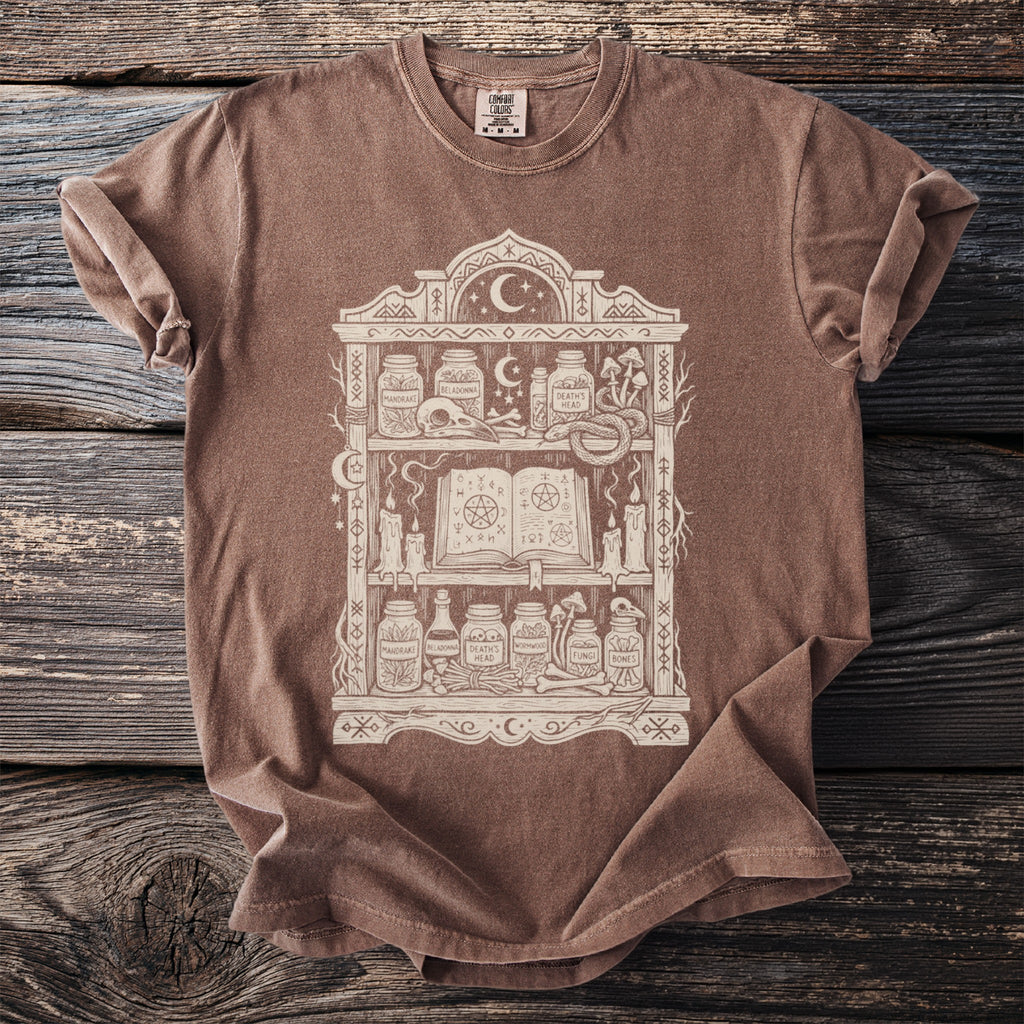 Brown t-shirt with a cream graphic of an occult shelf filled with jars, a skull, and a spellbook.