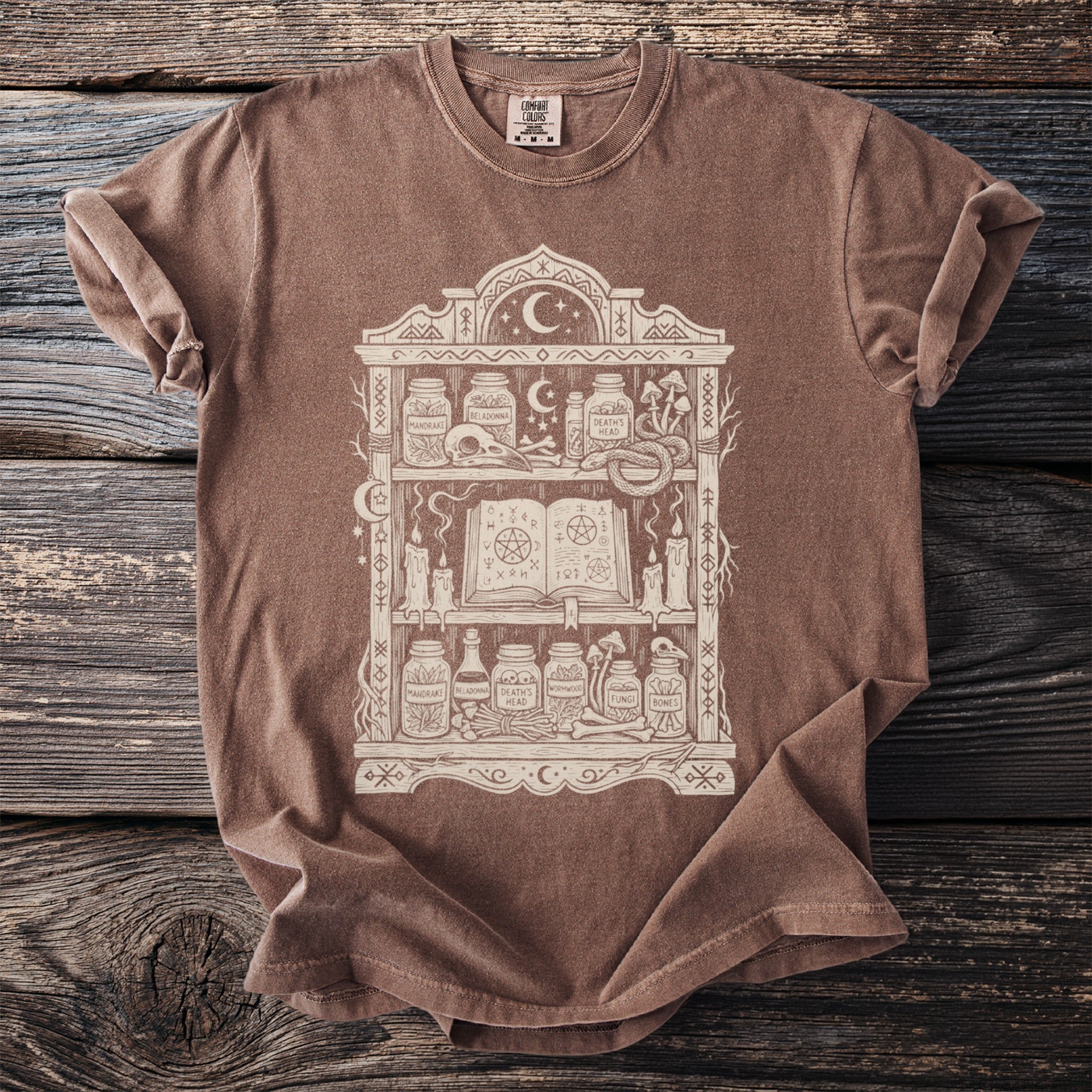 Brown t-shirt with a cream graphic of an occult shelf filled with jars, a skull, and a spellbook.