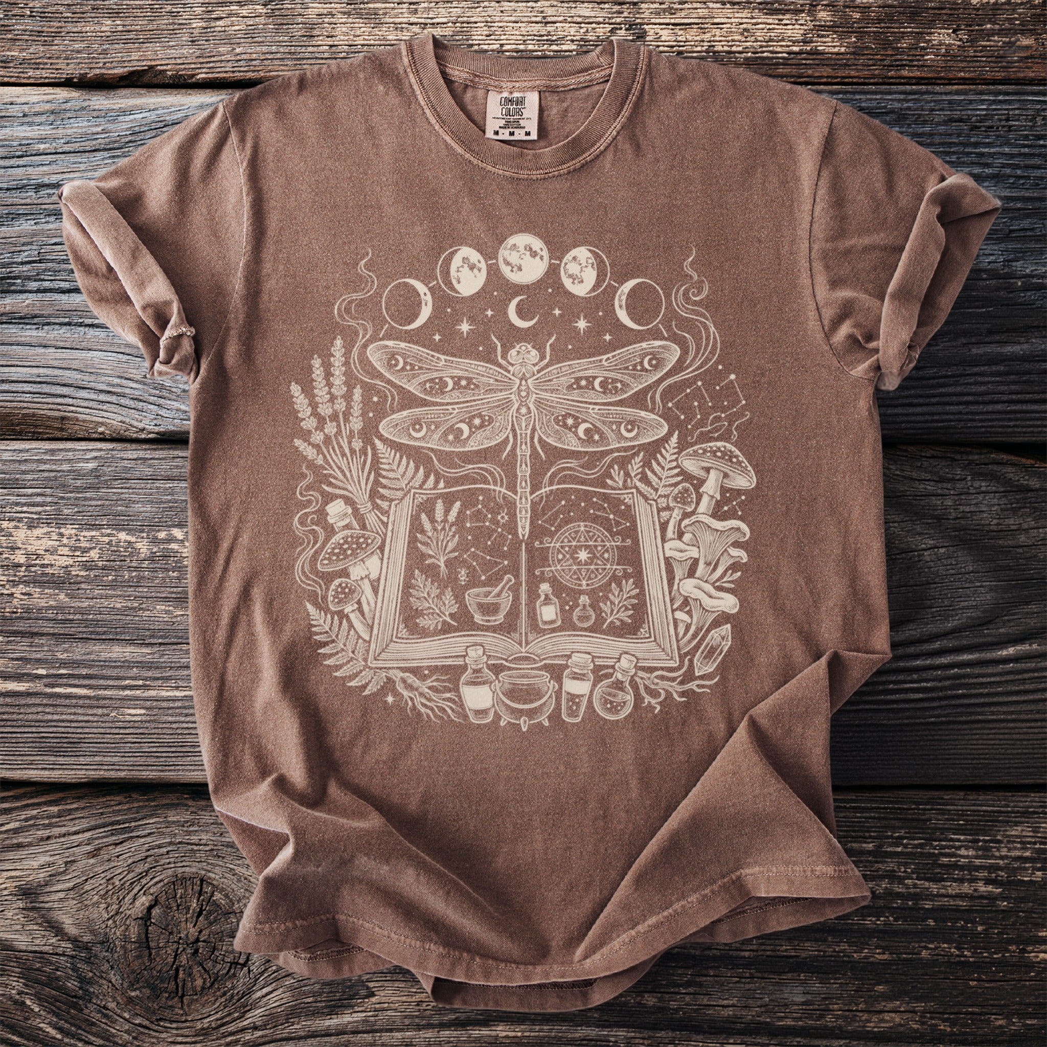 Brown t-shirt with white mystical dragonfly, moon phases, and potion ingredients graphic.