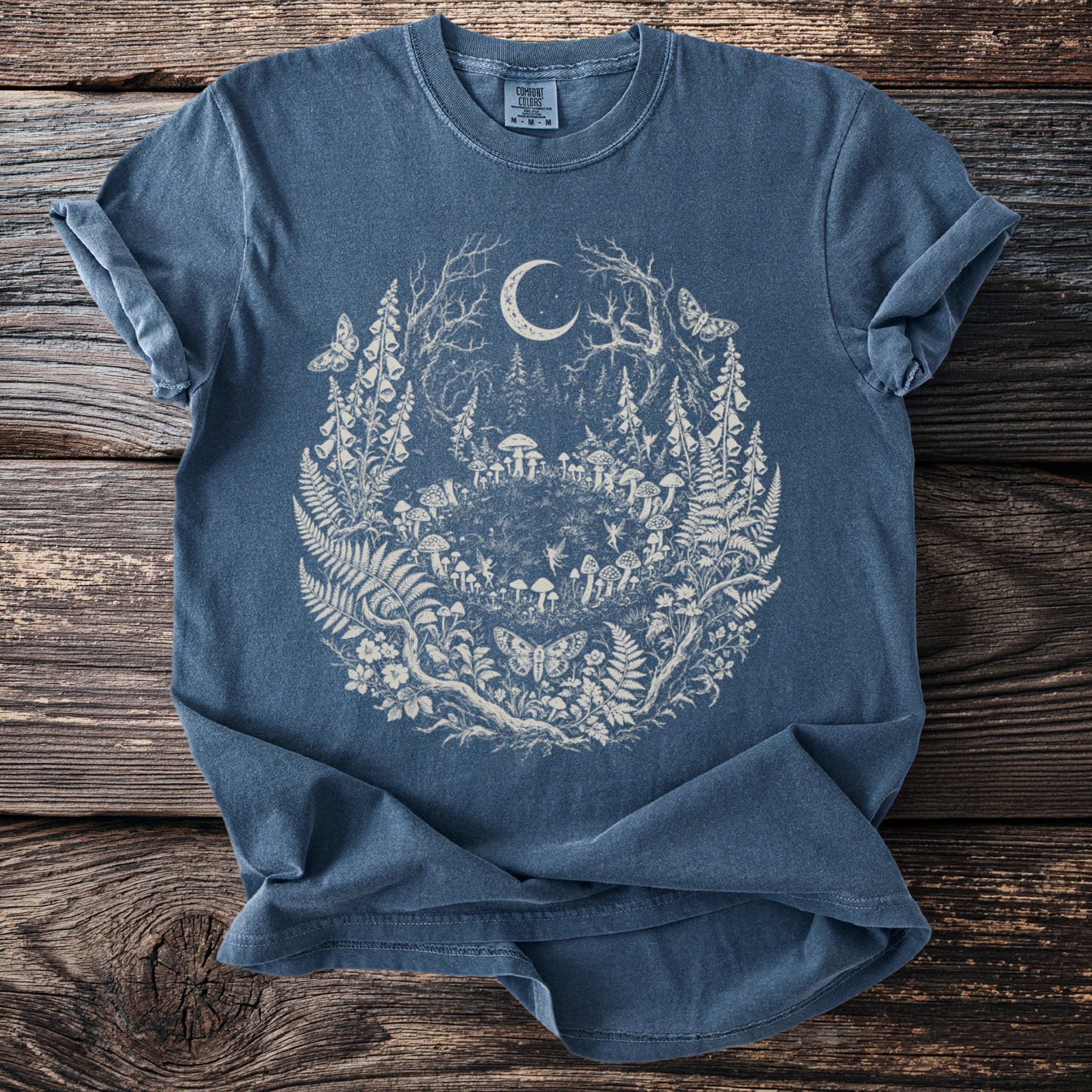 Blue t-shirt with a circular forest design featuring a crescent moon, mushrooms, fairies, and butterflies.