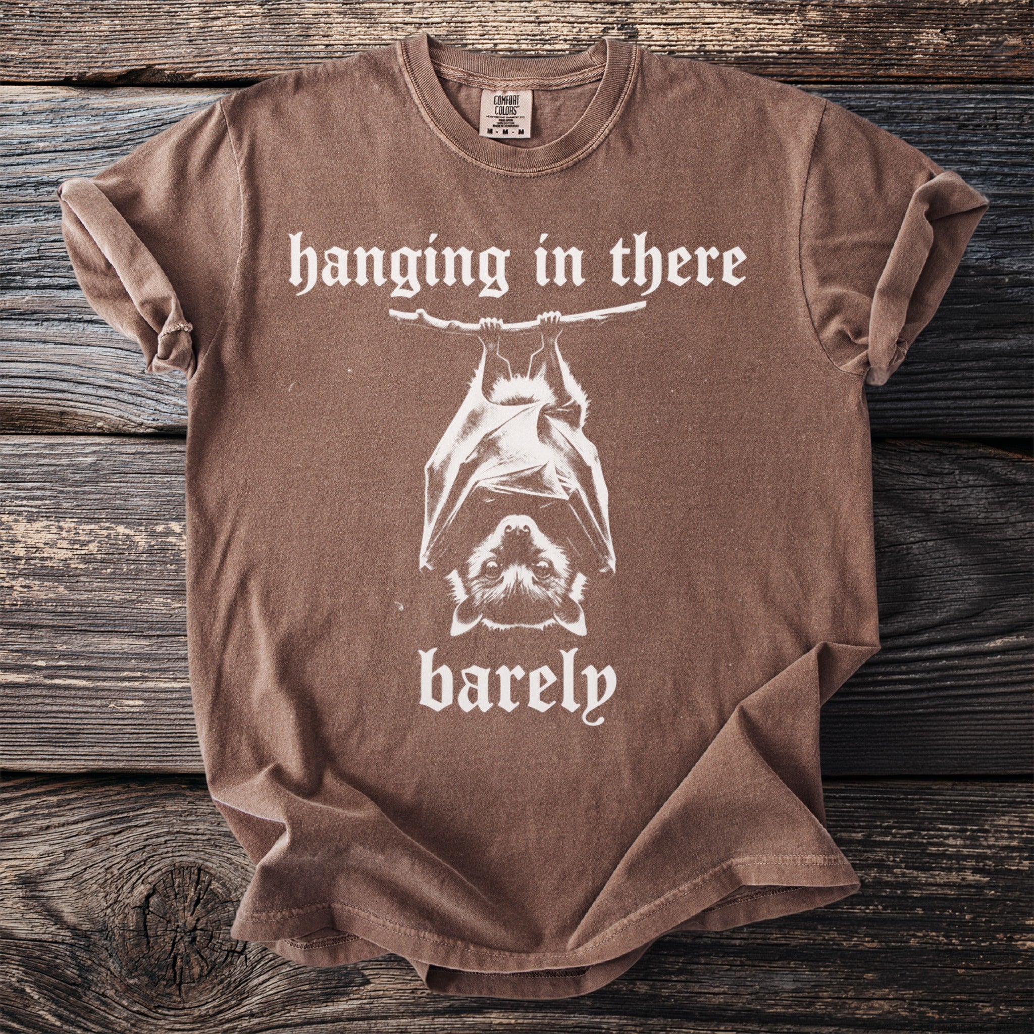 Brown t-shirt with a graphic of a bat hanging upside down and text "hanging in there barely".