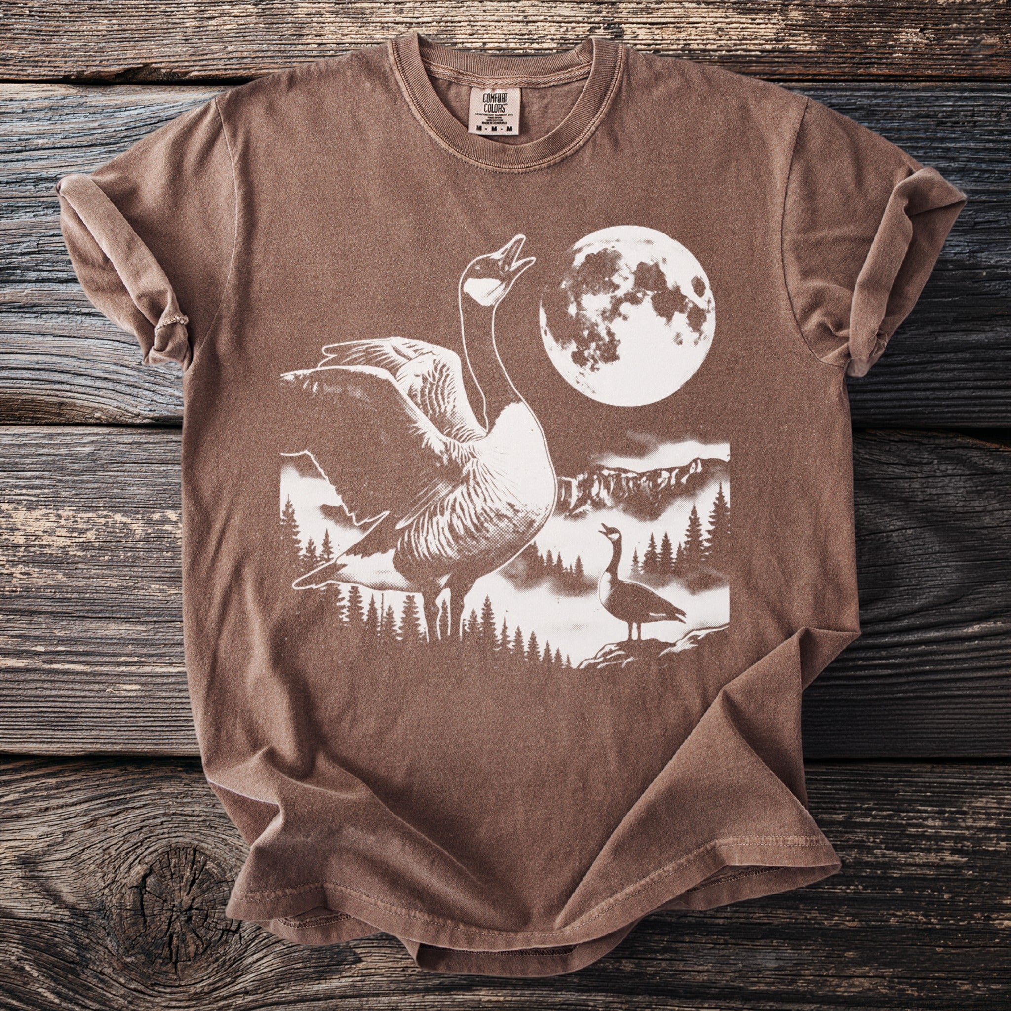 Brown t-shirt with a graphic of geese, mountains, trees, and a full moon.