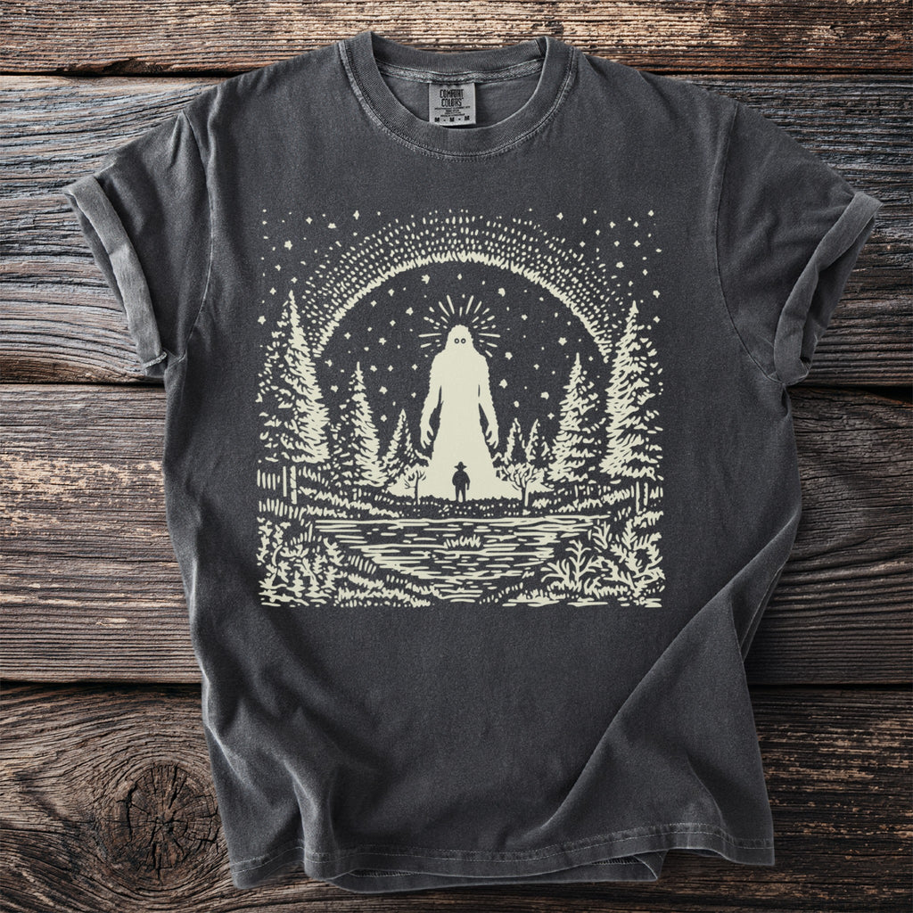 Dark grey t-shirt with a cream graphic of a cryptid in a forest beside a lake.