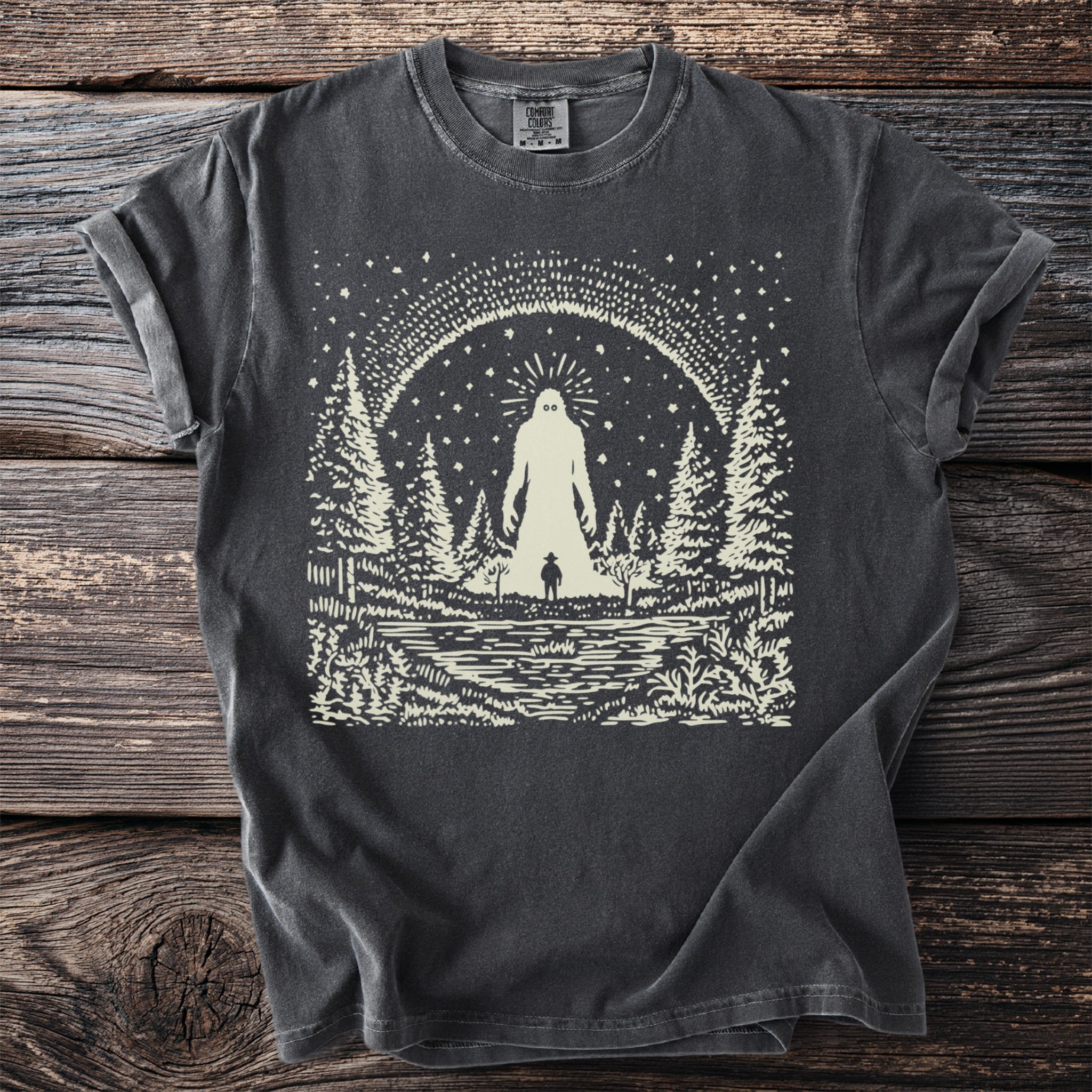 Dark grey t-shirt with a cream graphic of a cryptid in a forest beside a lake.
