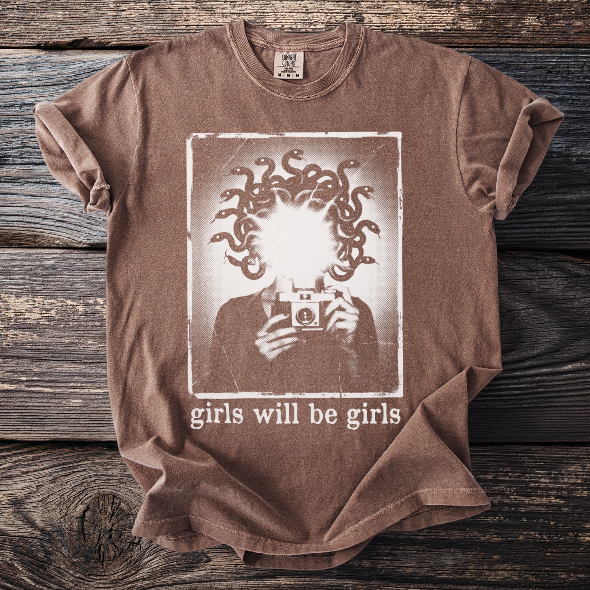 Brown t-shirt with graphic of person holding camera and snakes for hair, text "girls will be girls".