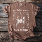 Brown t-shirt with graphic of three witches, a lantern, broom, and moon. Text reads "god forbid women have hobbies".
