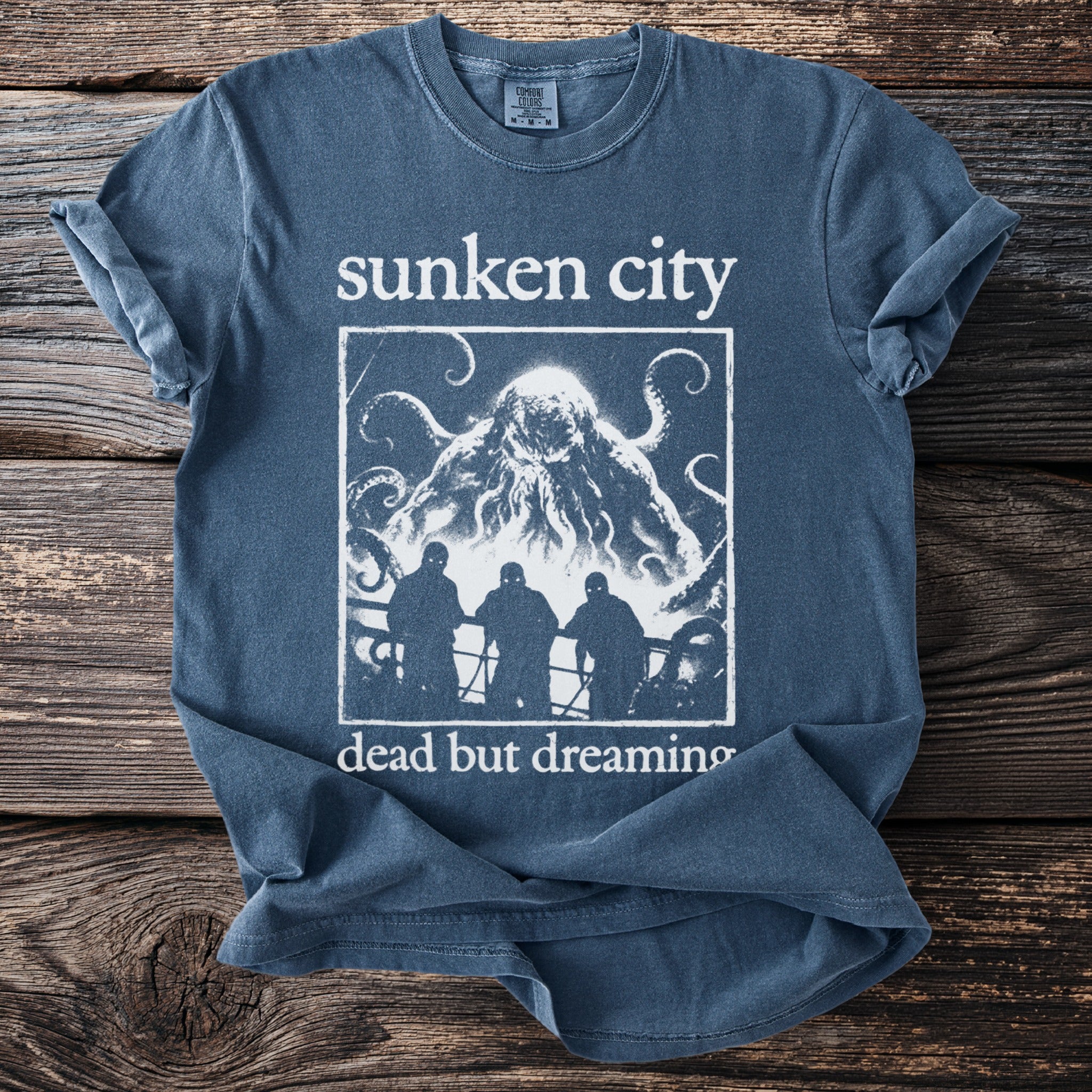 Blue t-shirt with "Sunken City" and "dead but dreaming" text and a creature graphic.