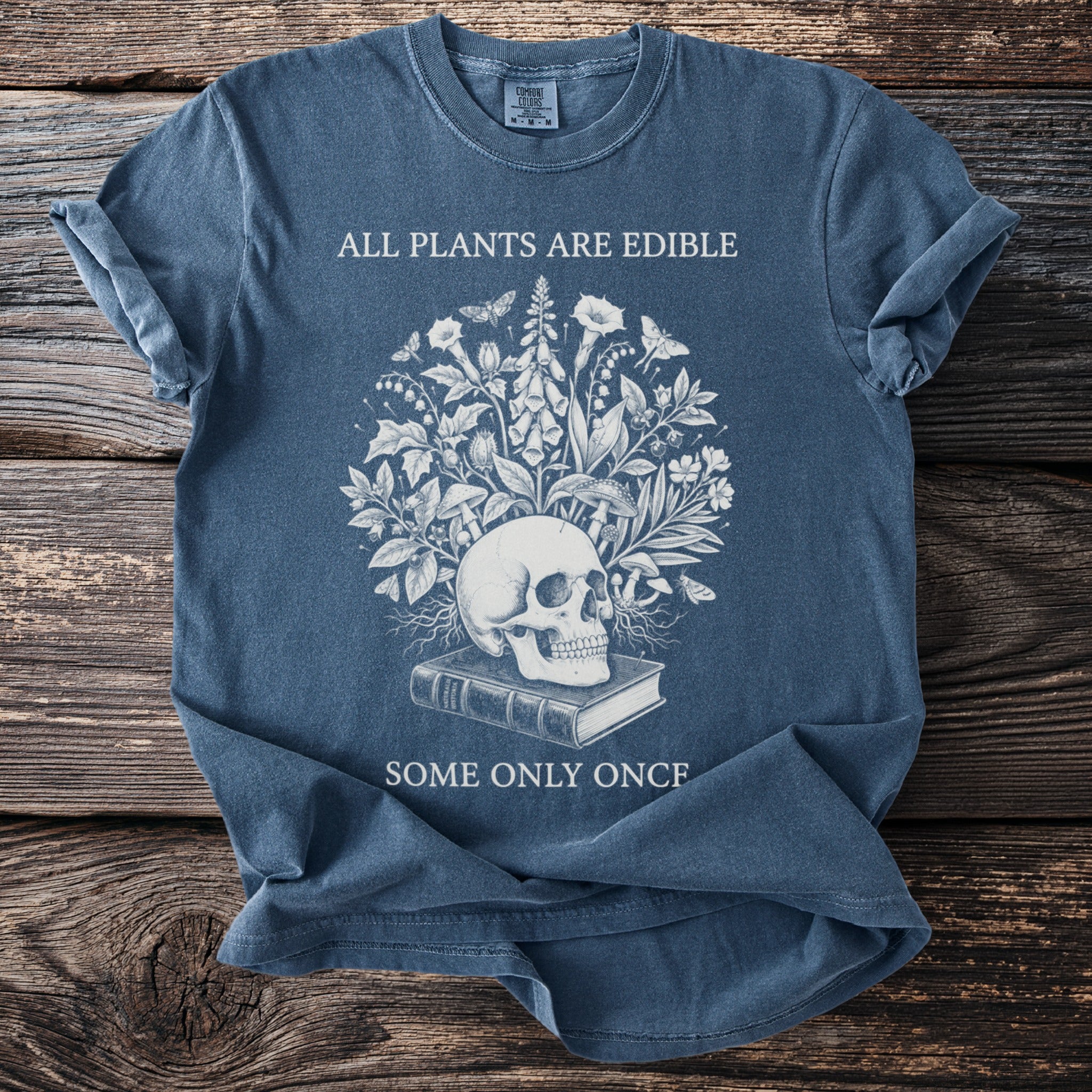 T-shirt with skull, book, plants, and text "ALL PLANTS ARE EDIBLE SOME ONLY ONCF".