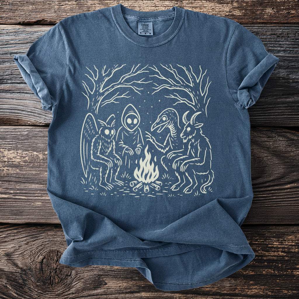 Blue t-shirt with cream illustration of mythical creatures around a campfire.