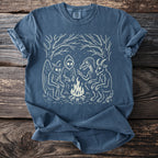Blue t-shirt with cream illustration of mythical creatures around a campfire.