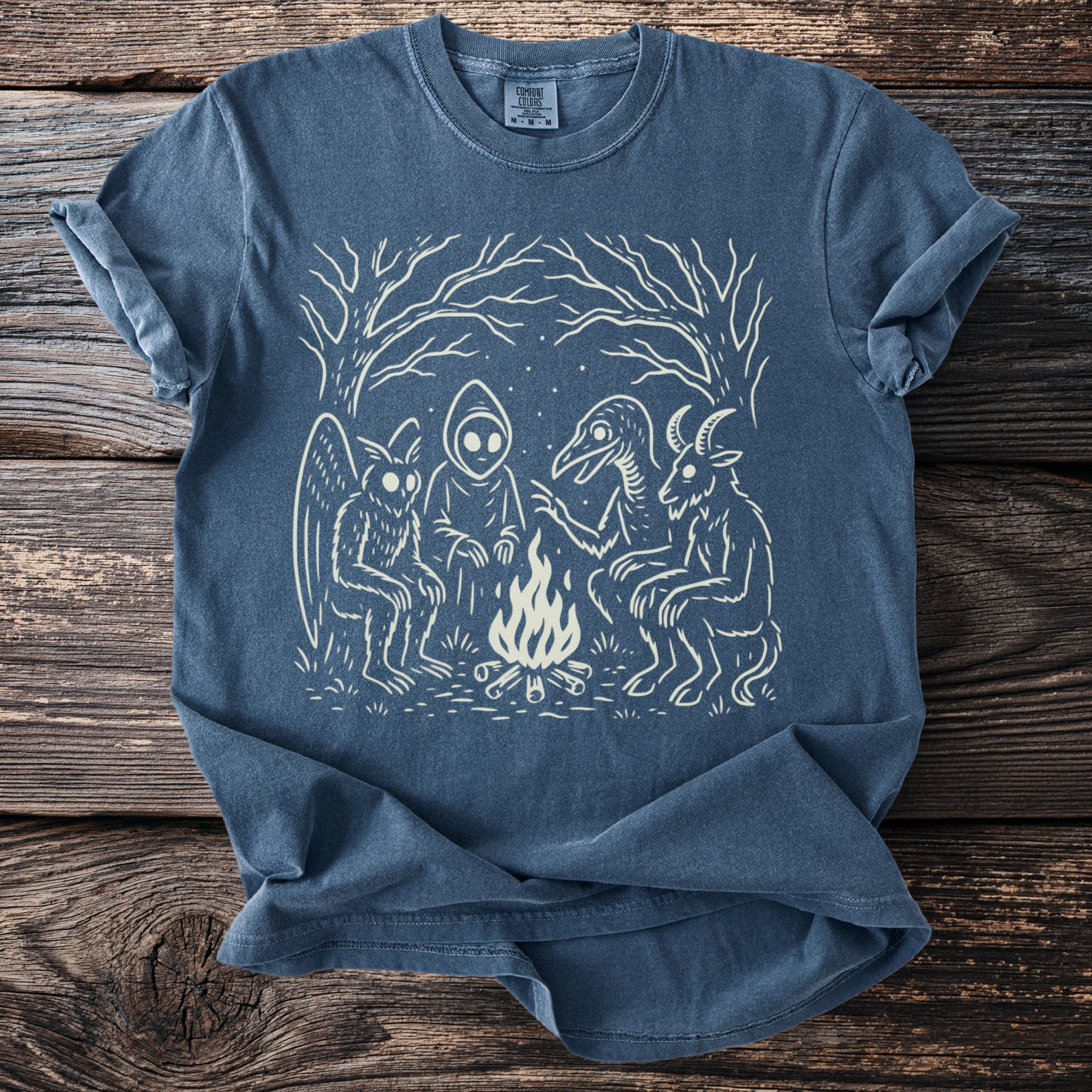 Blue t-shirt with cream illustration of mythical creatures around a campfire.