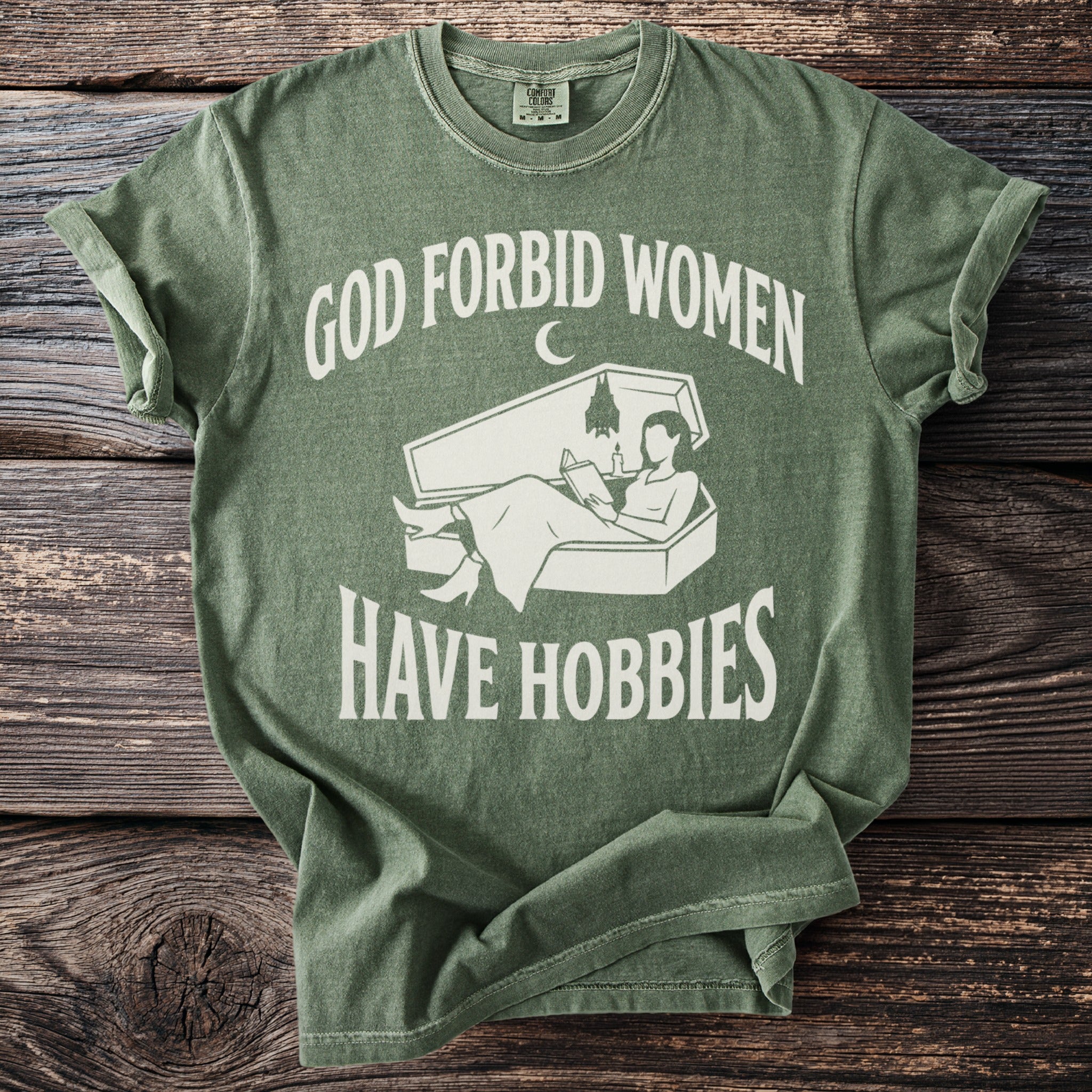 Green t-shirt with white text "God forbid women have hobbies" and coffin design.