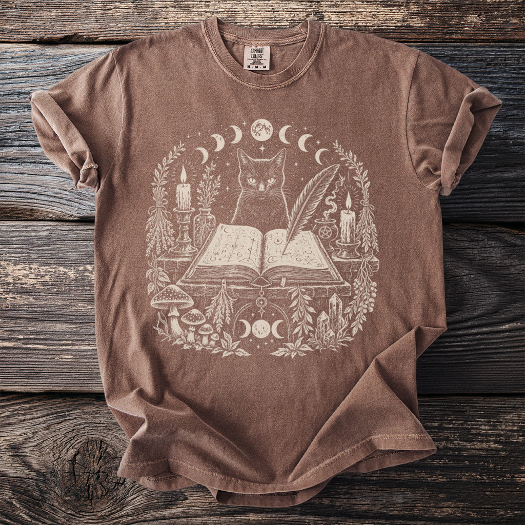 T-shirt with mystical cat, open book, candles, and celestial motifs in white print.