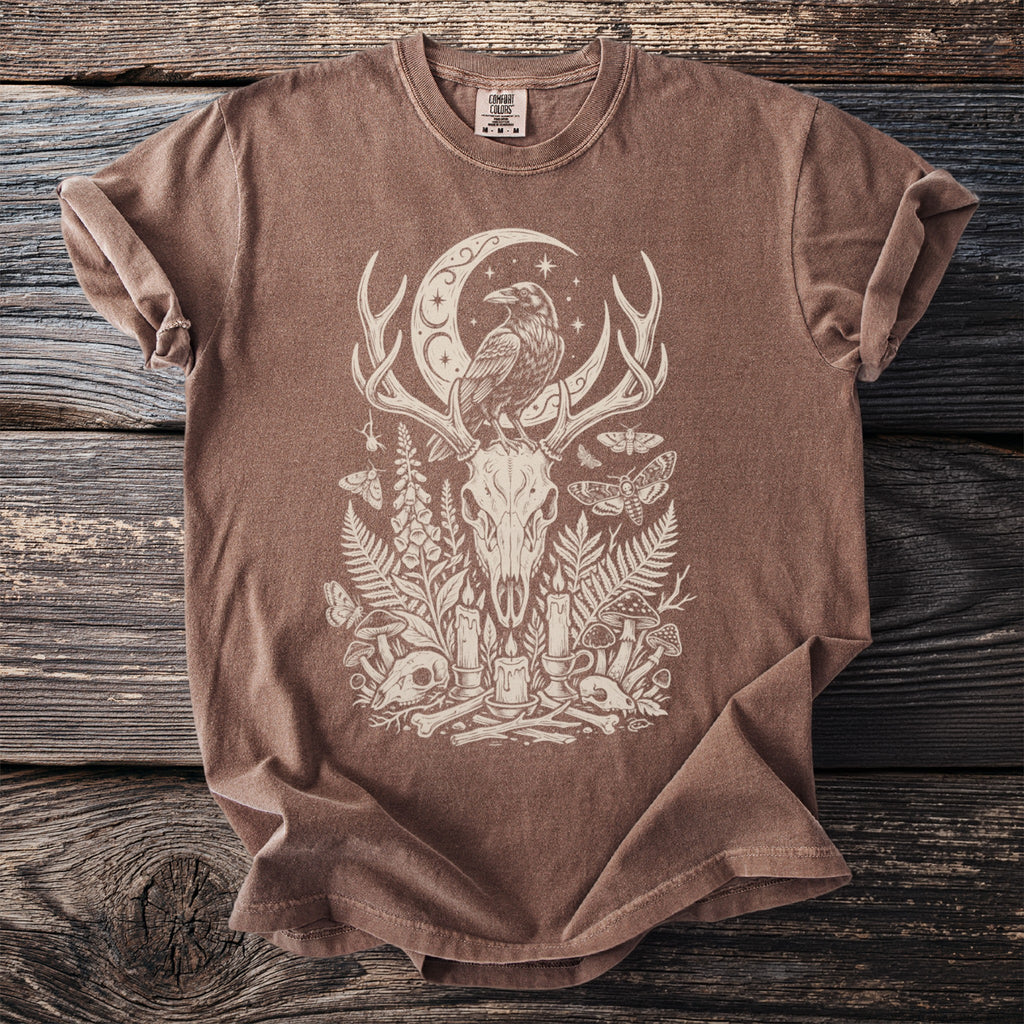 Brown t-shirt with a cream graphic depicting a raven on a crescent moon above a deer skull.