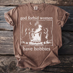 Brown t-shirt with white graphic of witches dancing around a fire and text "god forbid women have hobbies".