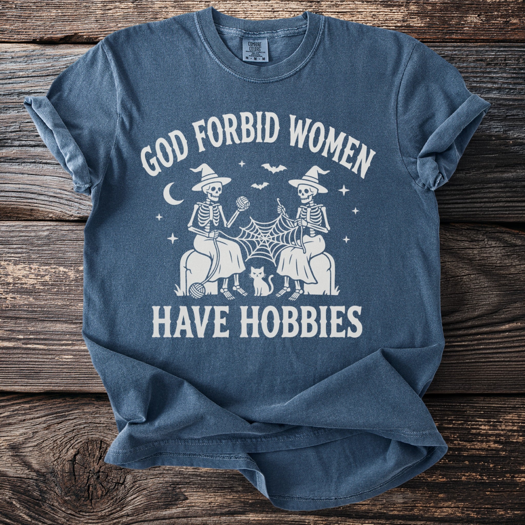 Blue t-shirt with white text and graphics of knitting skeletons and a cat.