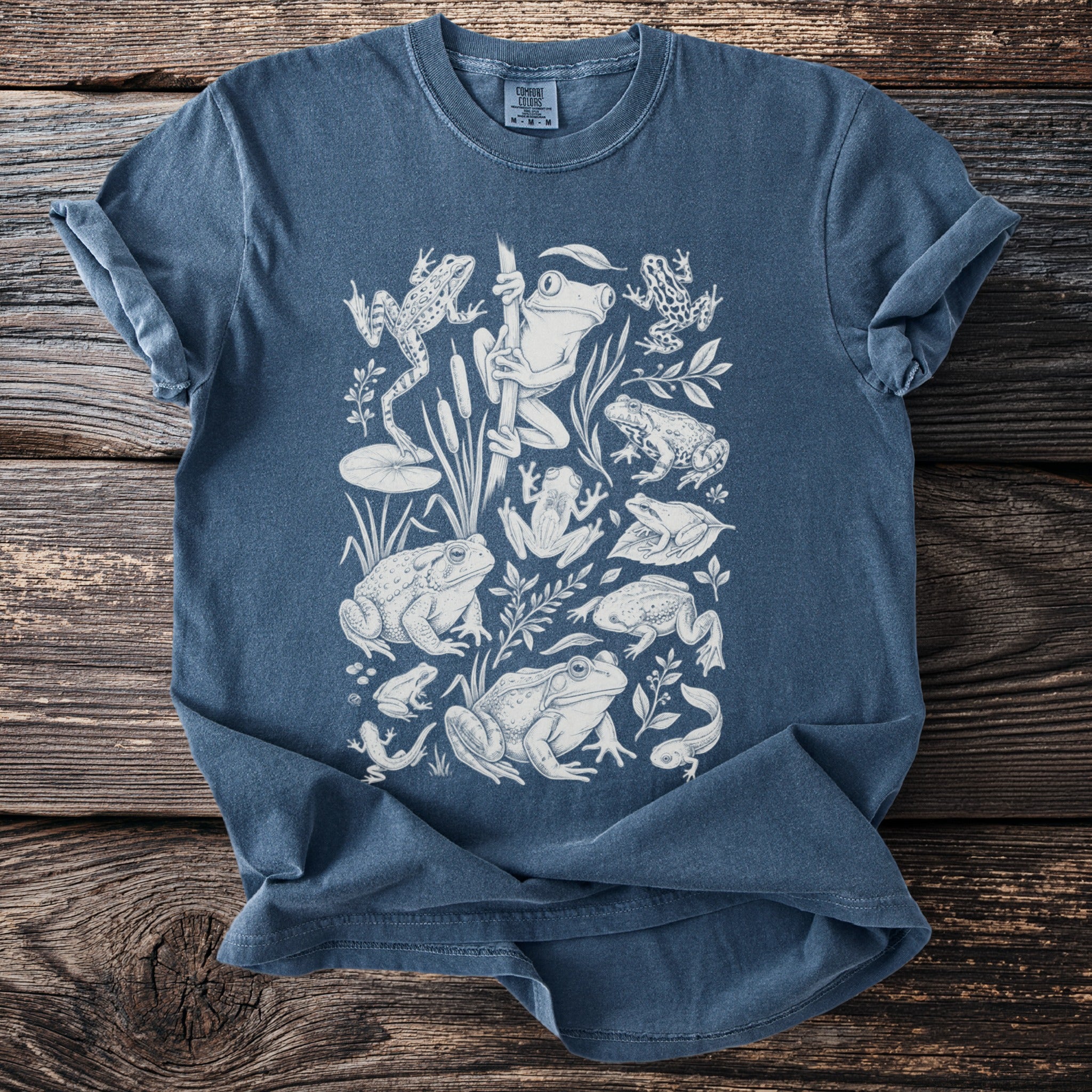 Blue t-shirt with white line art illustration of various frogs and tadpoles.
