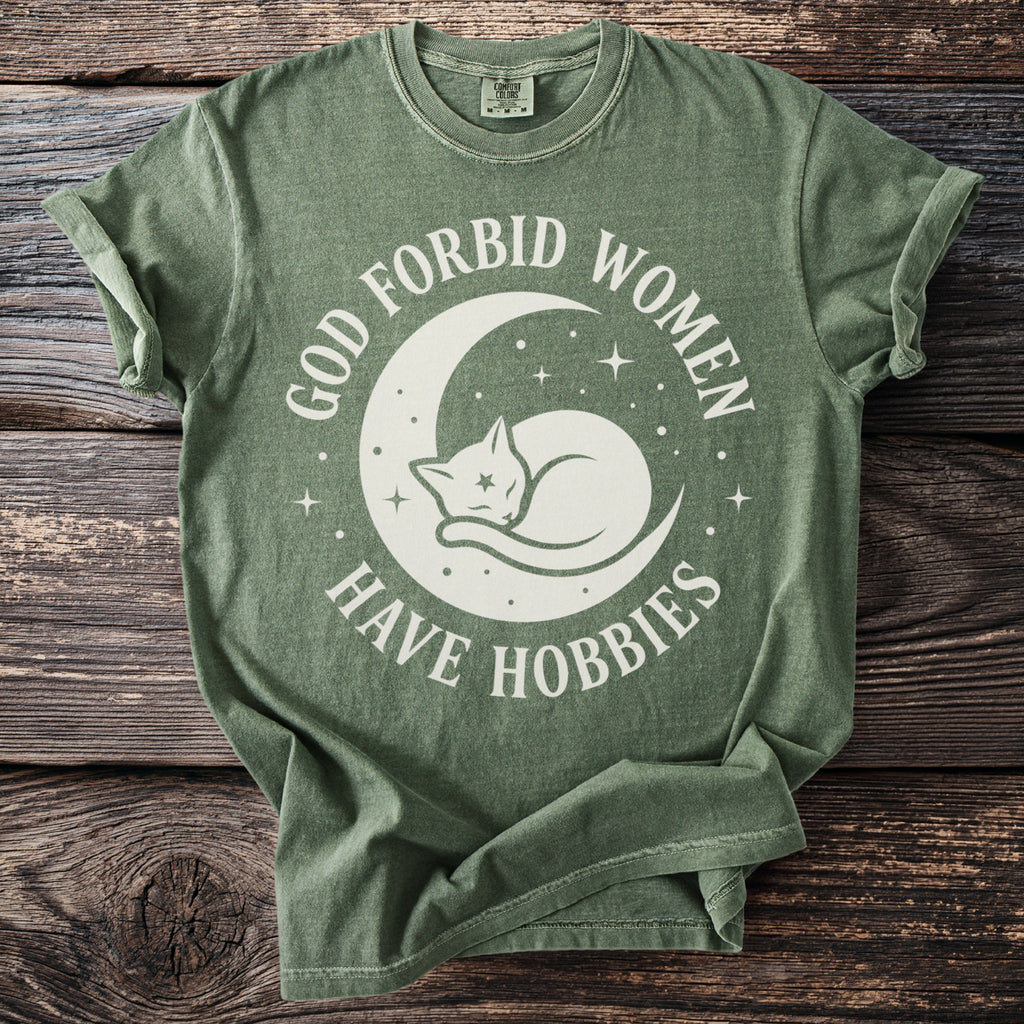 Green t-shirt with white graphic of a cat on a crescent moon and text "God forbid women have hobbies".
