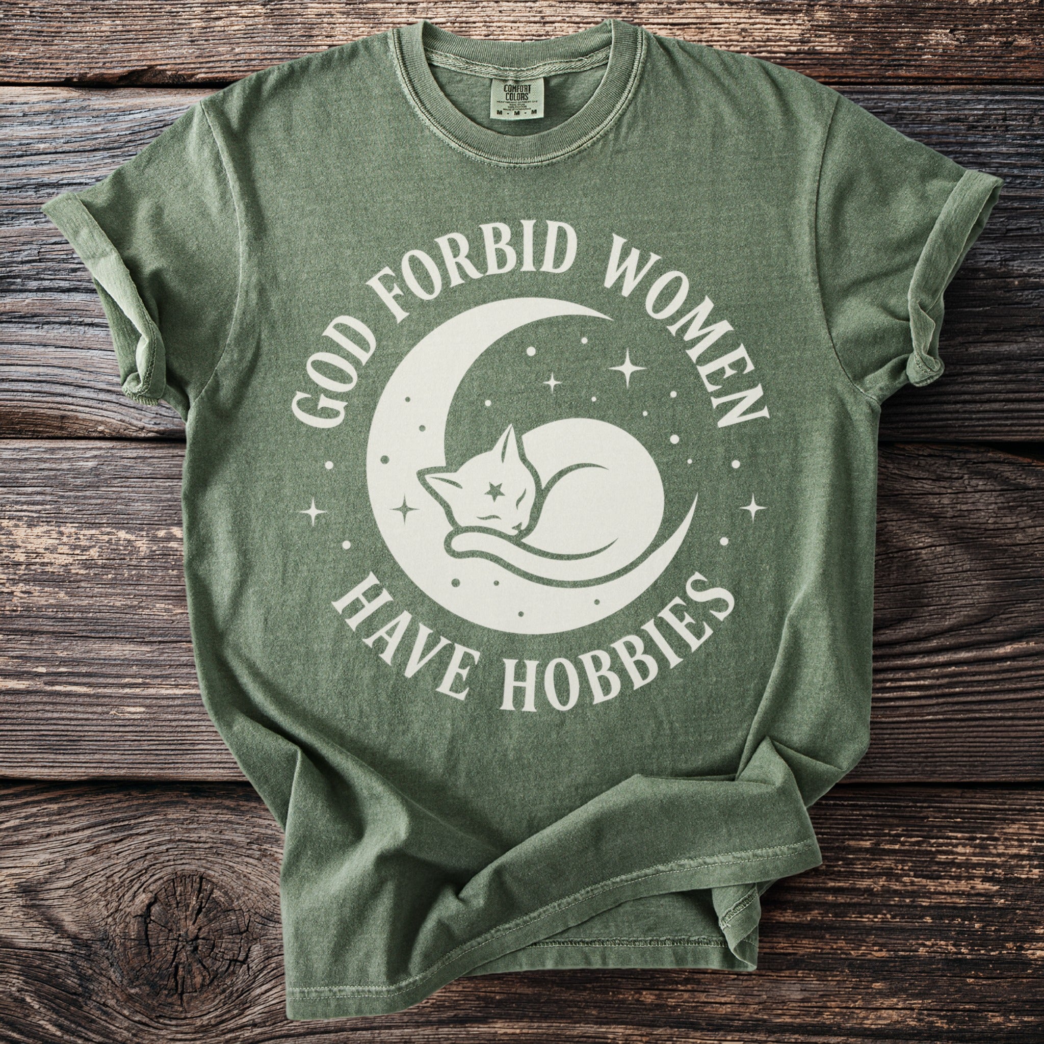 Green t-shirt with white graphic of a cat on a crescent moon and text "God forbid women have hobbies".