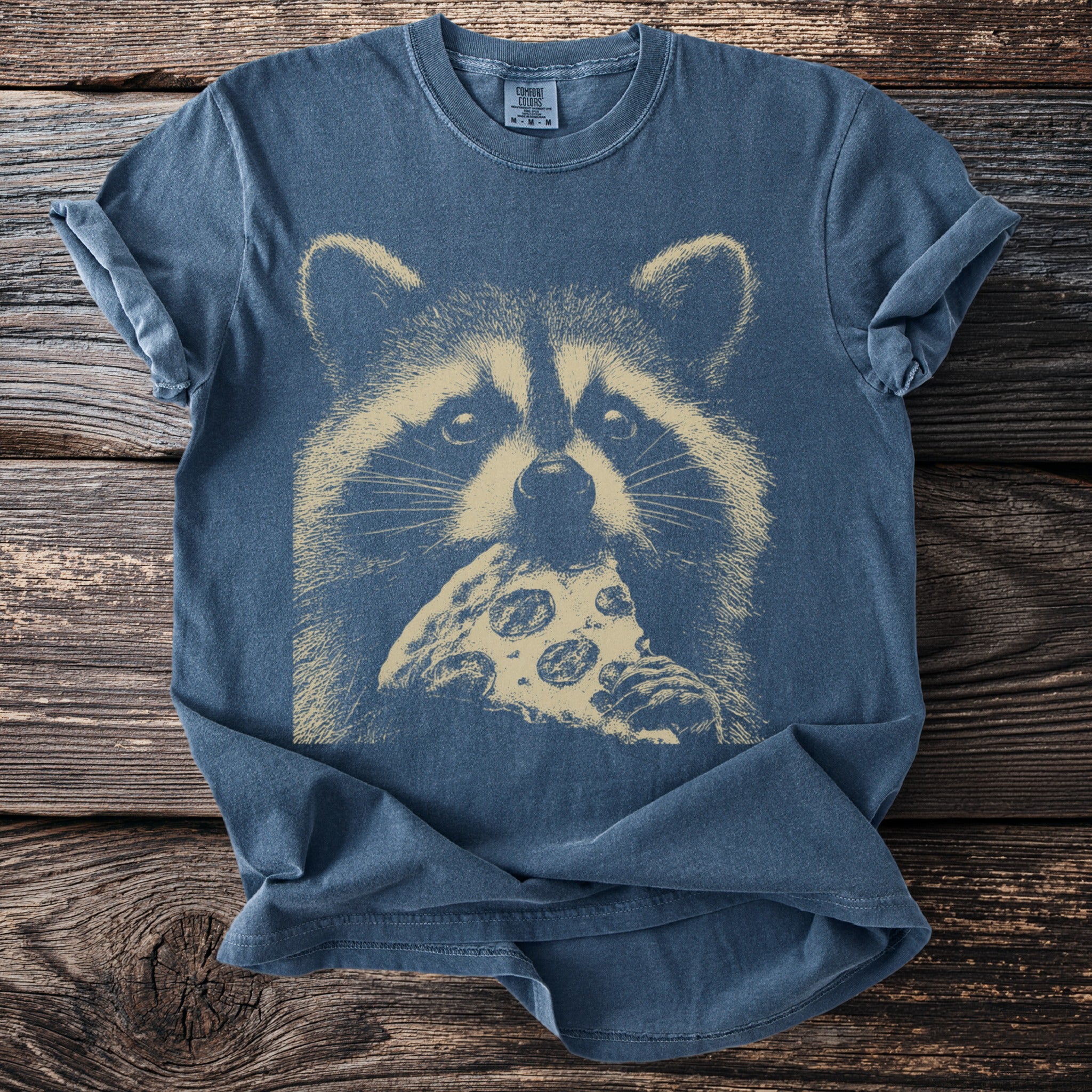 Blue t-shirt with a beige graphic of a raccoon eating pizza.
