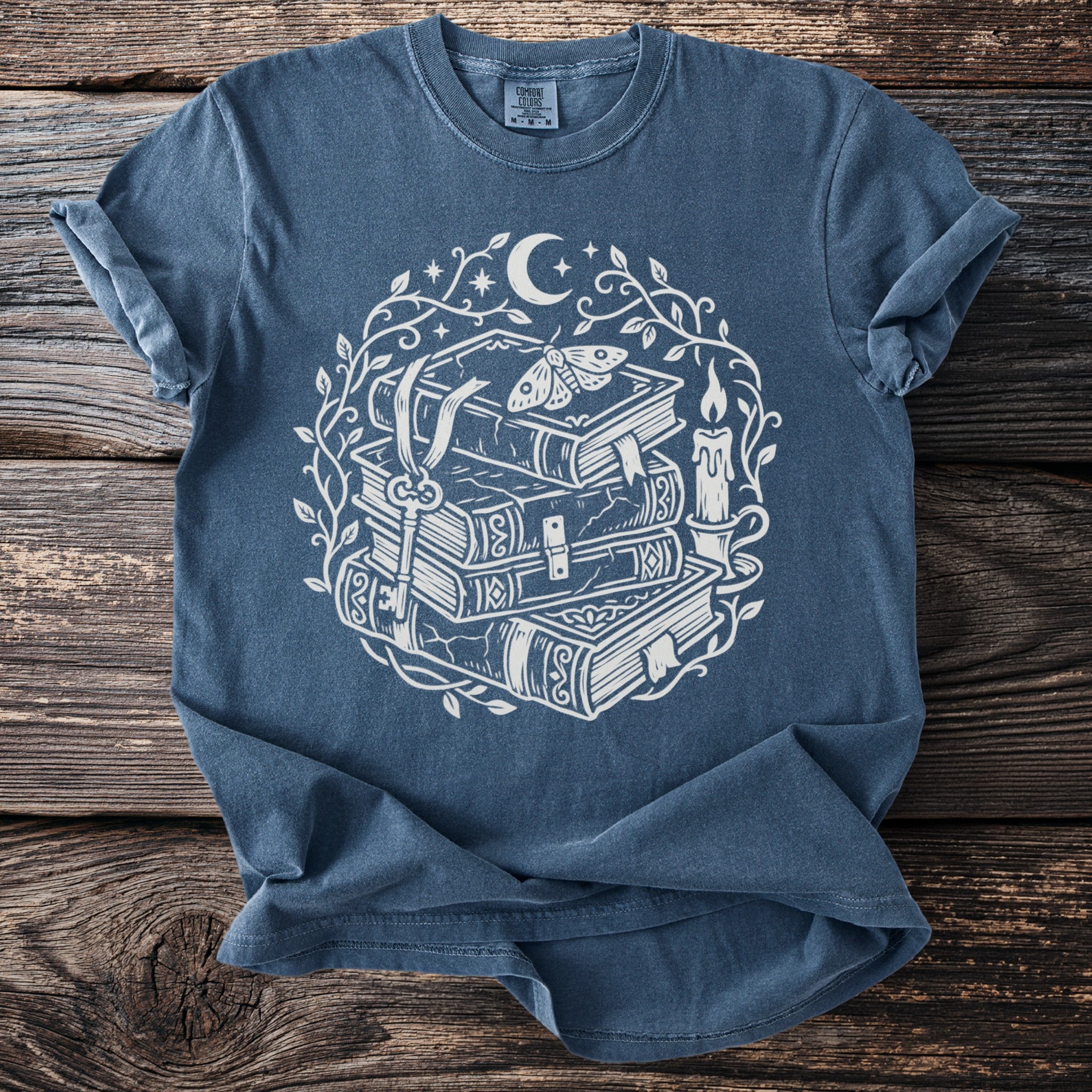 Blue t-shirt with white illustration of stacked books, key, moth, crescent moon, and candle.