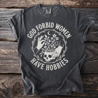 Dark gray t-shirt with white text "God forbid women have hobbies" and celestial skull graphic.