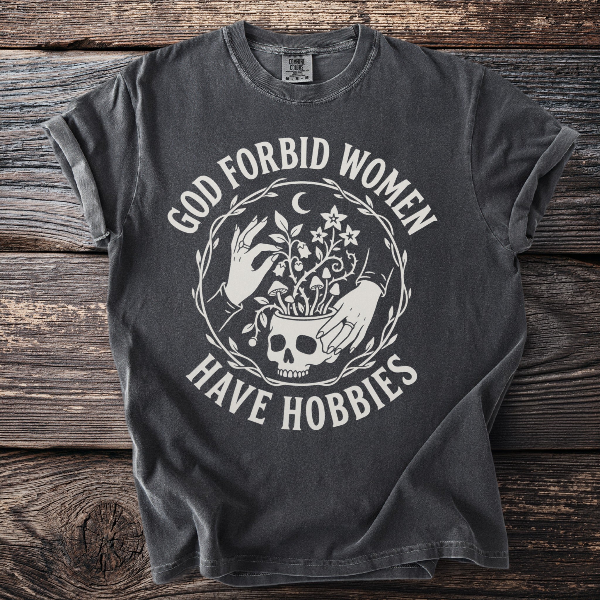 Dark gray t-shirt with white text "God forbid women have hobbies" and celestial skull graphic.