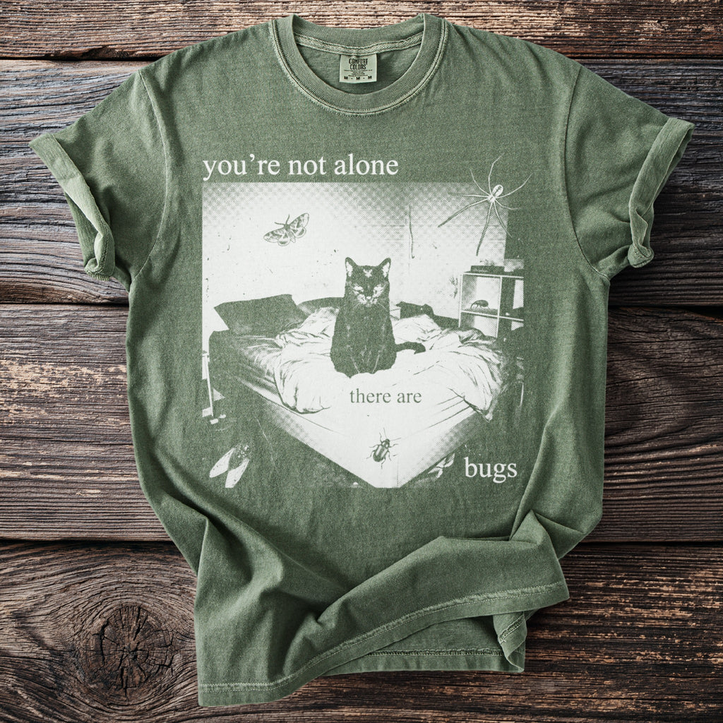 Green t-shirt with print: cat on bed, spider, moth, flies, and text "you're not alone there are bugs".