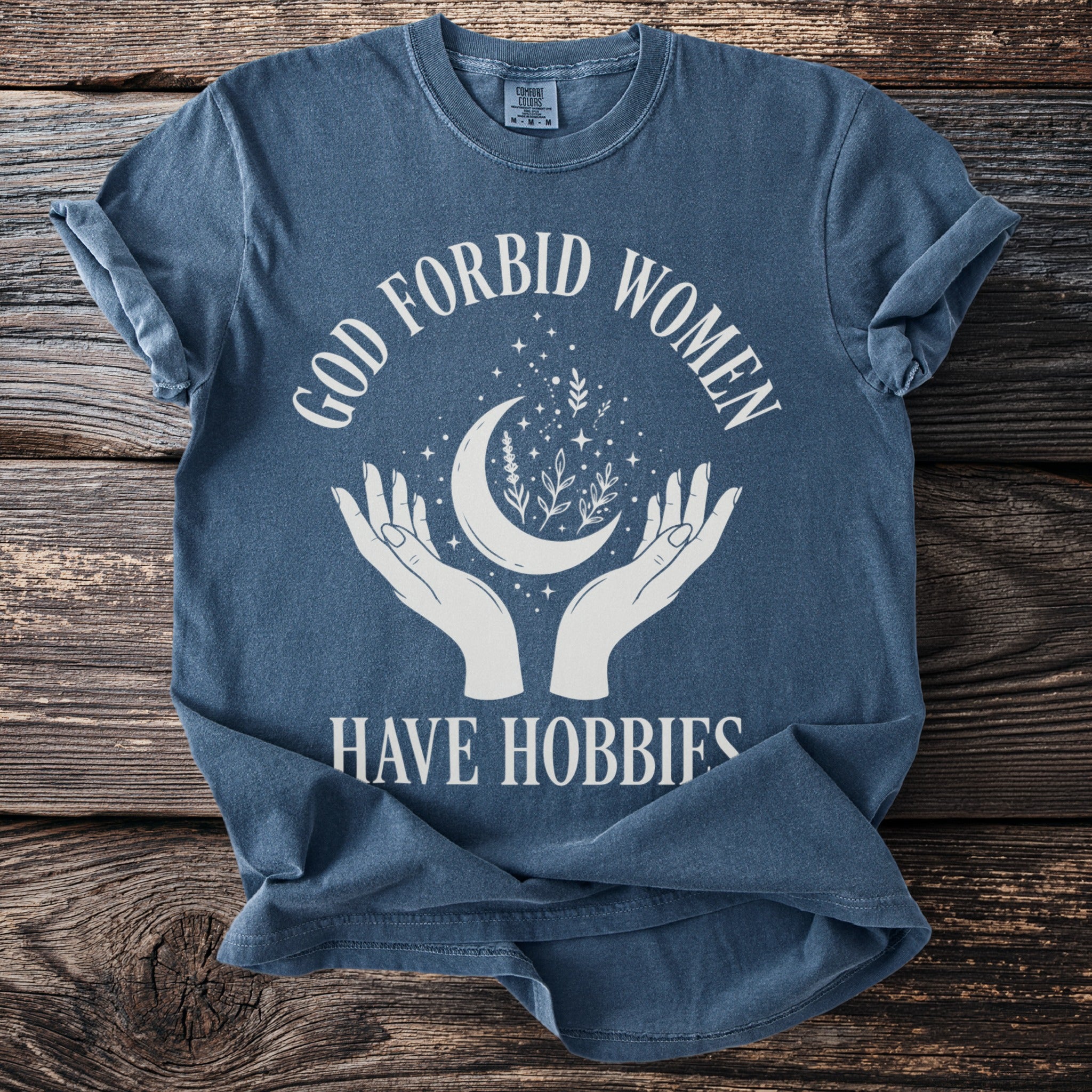 Blue t-shirt with text "GOD FORBID WOMEN HAVE HOBBIES" and celestial crescent moon graphic.