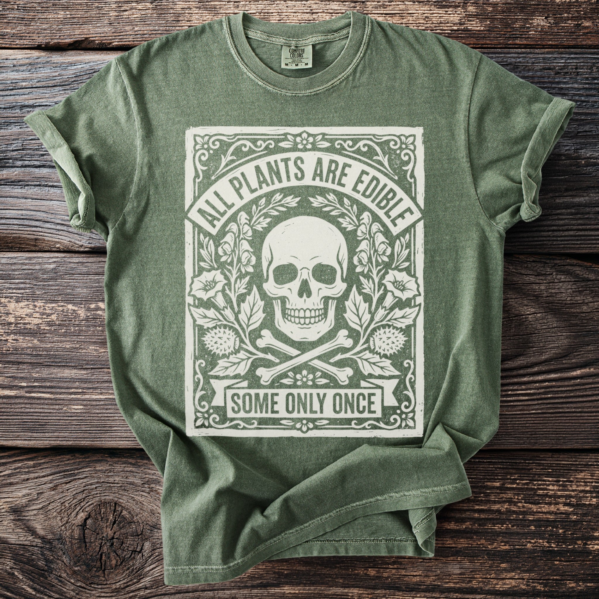 Green t-shirt with white graphic of a skull, bones, flowers, and text saying "ALL PLANTS ARE EDIBLE SOME ONLY ONCE".