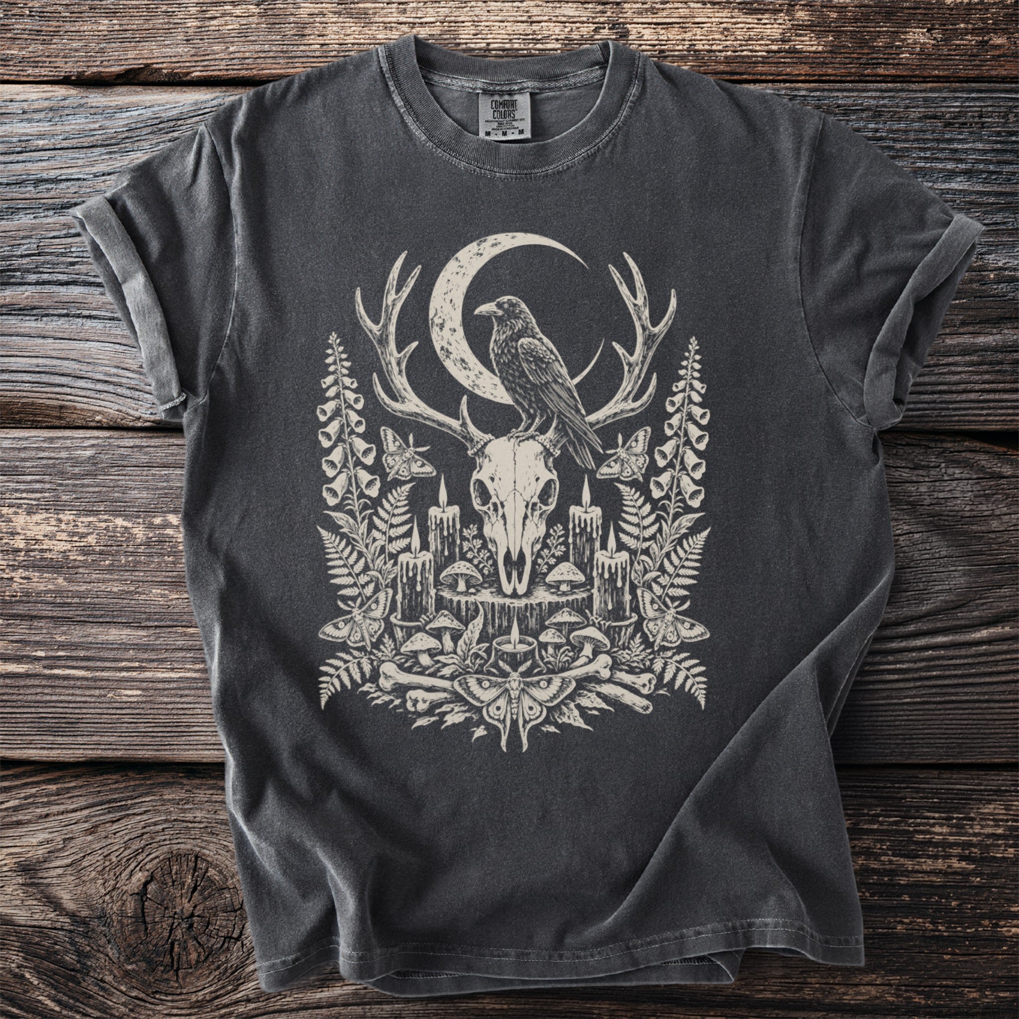 Dark t-shirt graphic of a raven on antlers above a skull with mushrooms and candles.