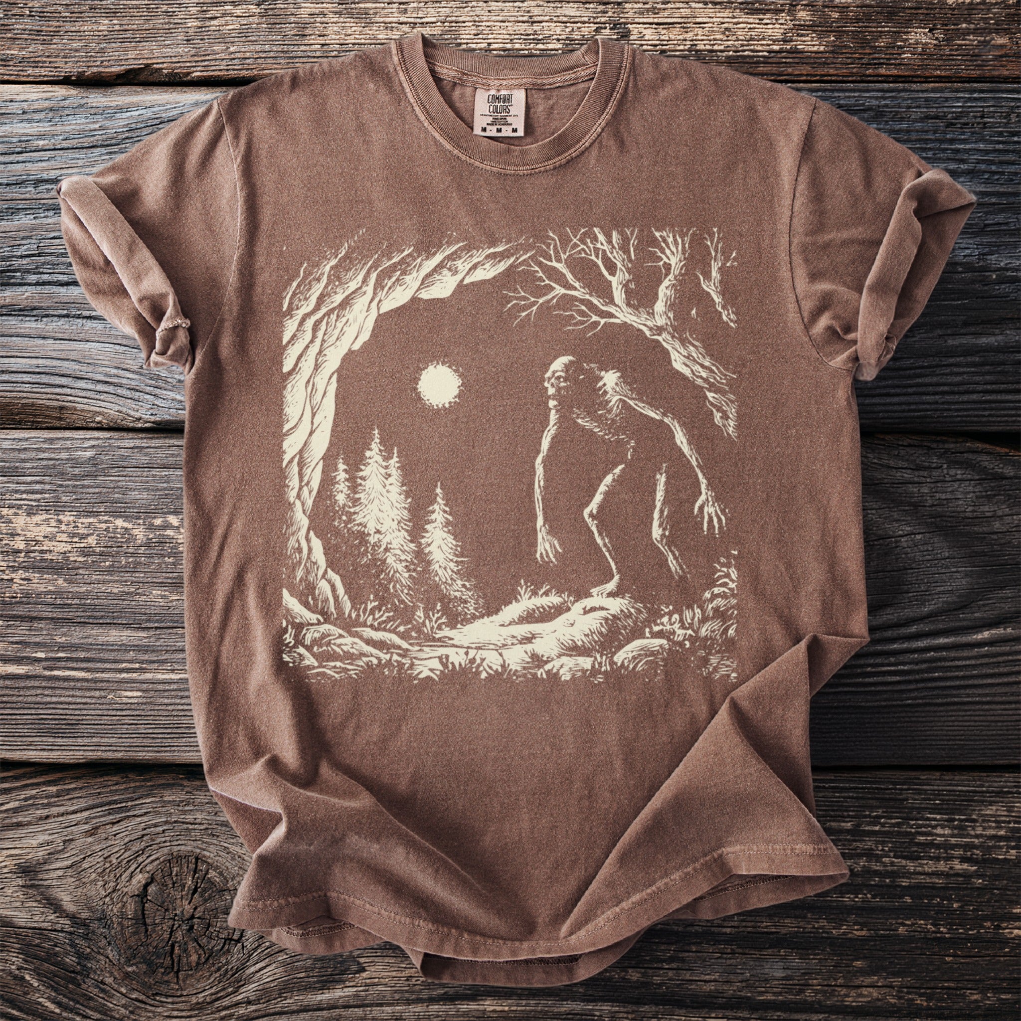Brown Comfort Colors t-shirt featuring a Bigfoot in a forest scene under a full moon.