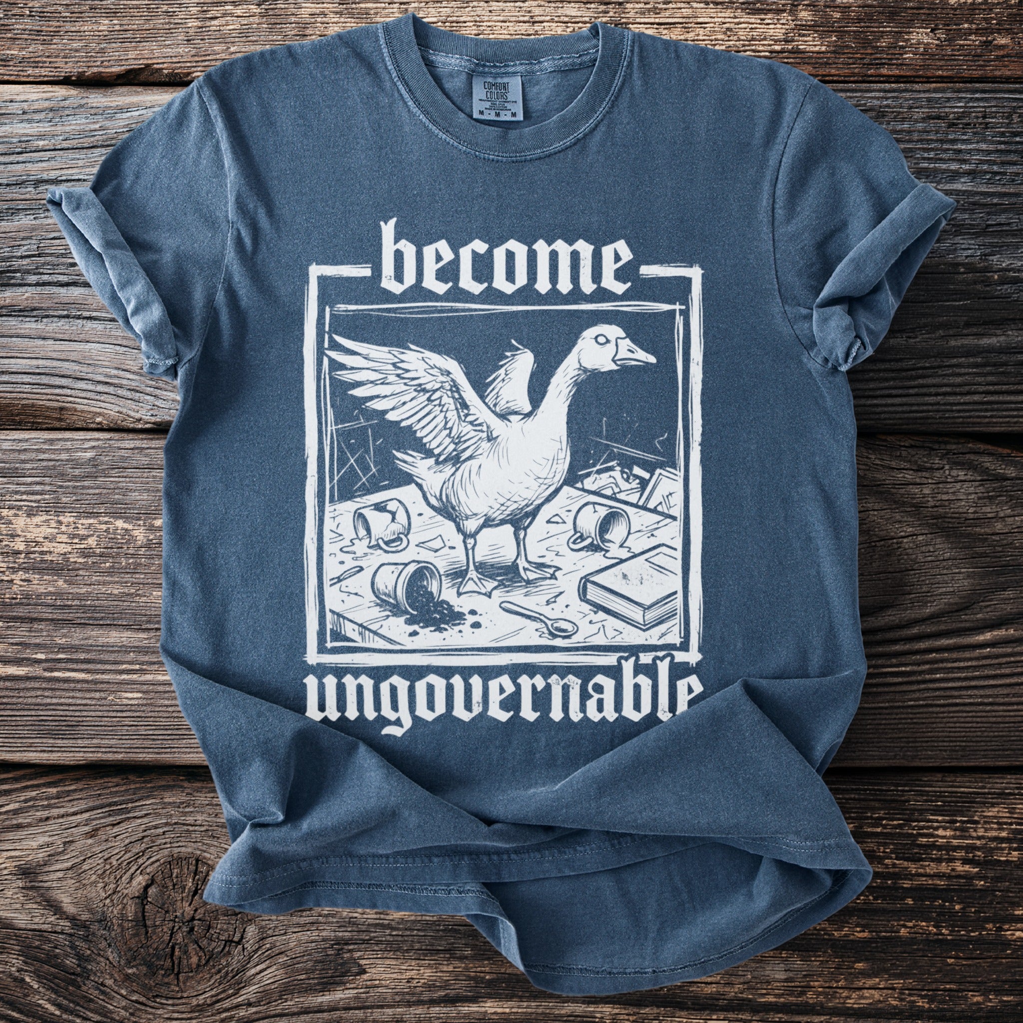 Blue t-shirt with white text "become" and a goose in a chaotic scene.