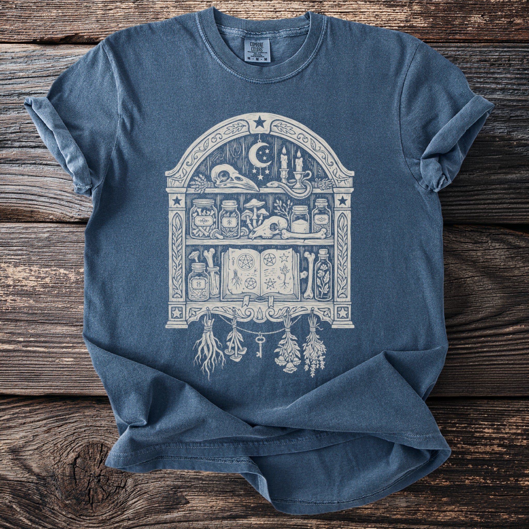 Blue t-shirt with an illustration of a witch's shelf filled with occult items.